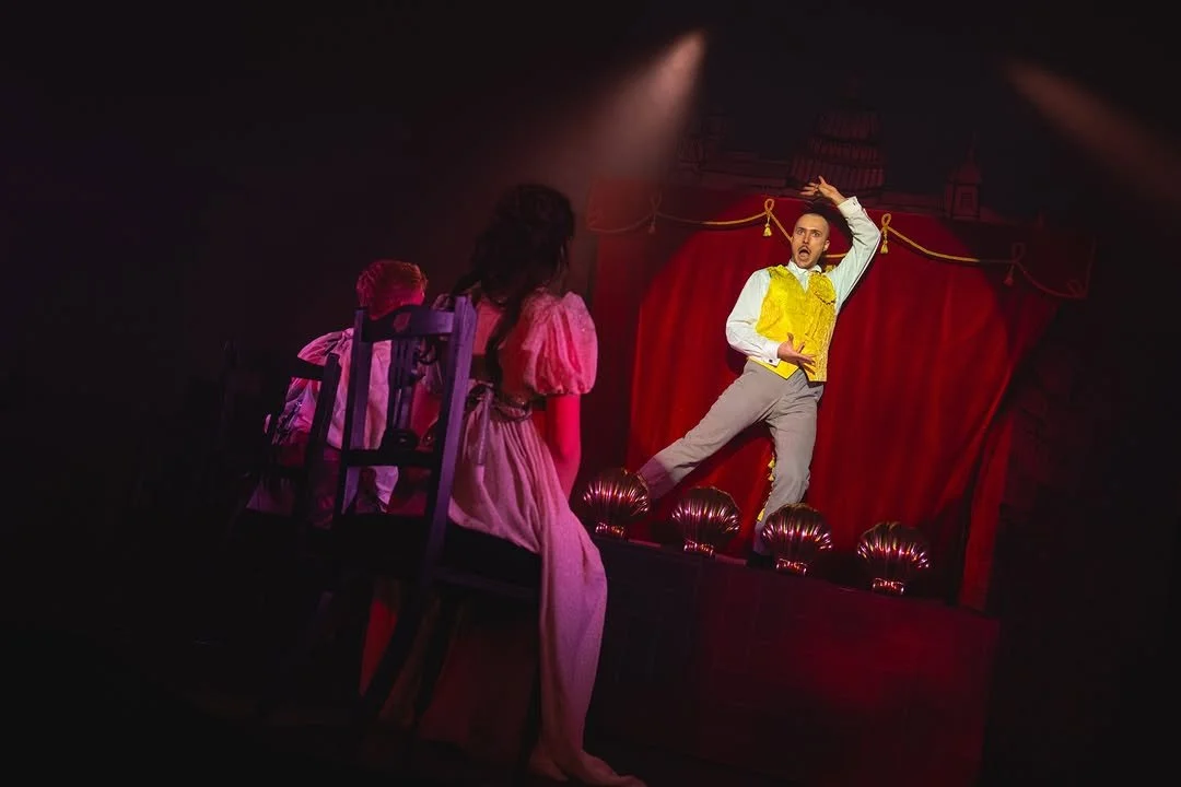 Actors perform on stage with red curtain backdrop, one man standing on a platform with a yellow vest, two women seated in front, stage lighting highlighting the scene.