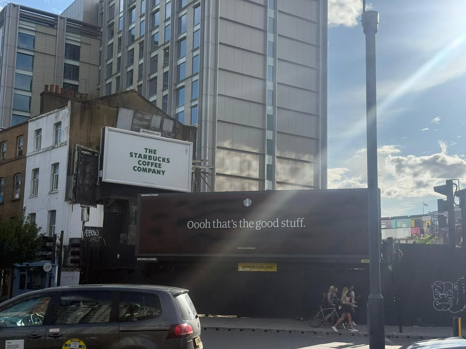 City street scene with a billboard for Starbucks Coffee Company, a digital billboard with the message 'Oooh that's the good stuff', multiple pedestrians, cars, and tall modern buildings in the background under a partly cloudy sky.