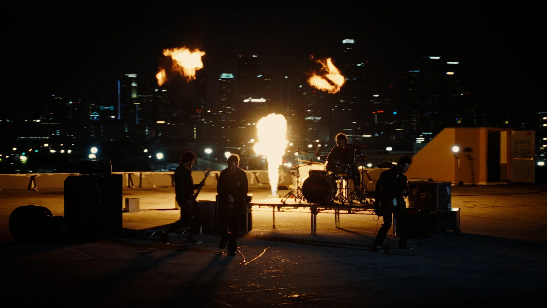 A band performing on a rooftop at night with city skyline in the background, pyrotechnics emitting flames behind them.