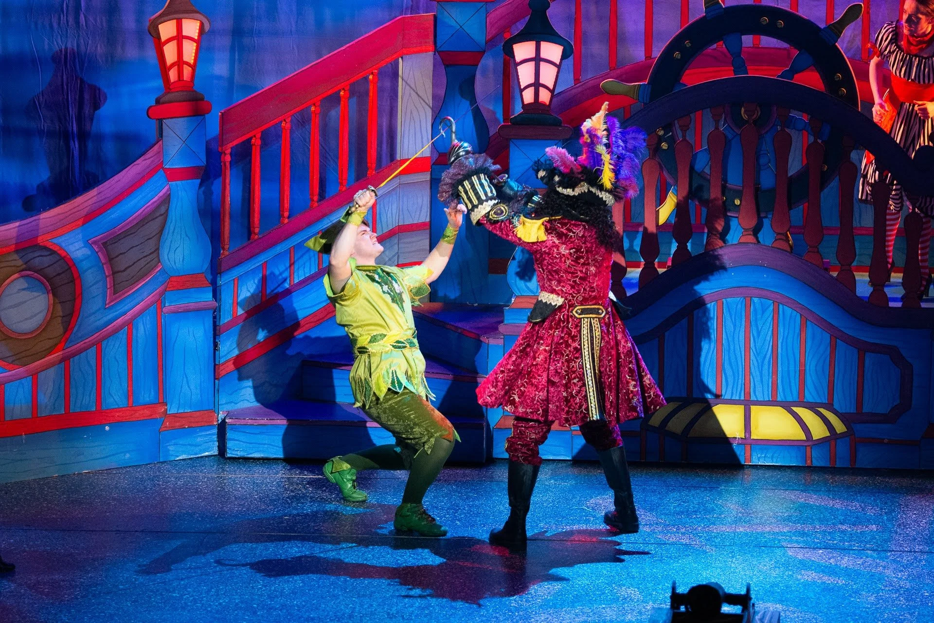 Two performers in colorful costumes act on a stage designed as a whimsical, vibrant pirate ship. One performer, in green and yellow, appears to be kneeling and holding a sword or stick, while the other, dressed in a red and black ornate costume with 