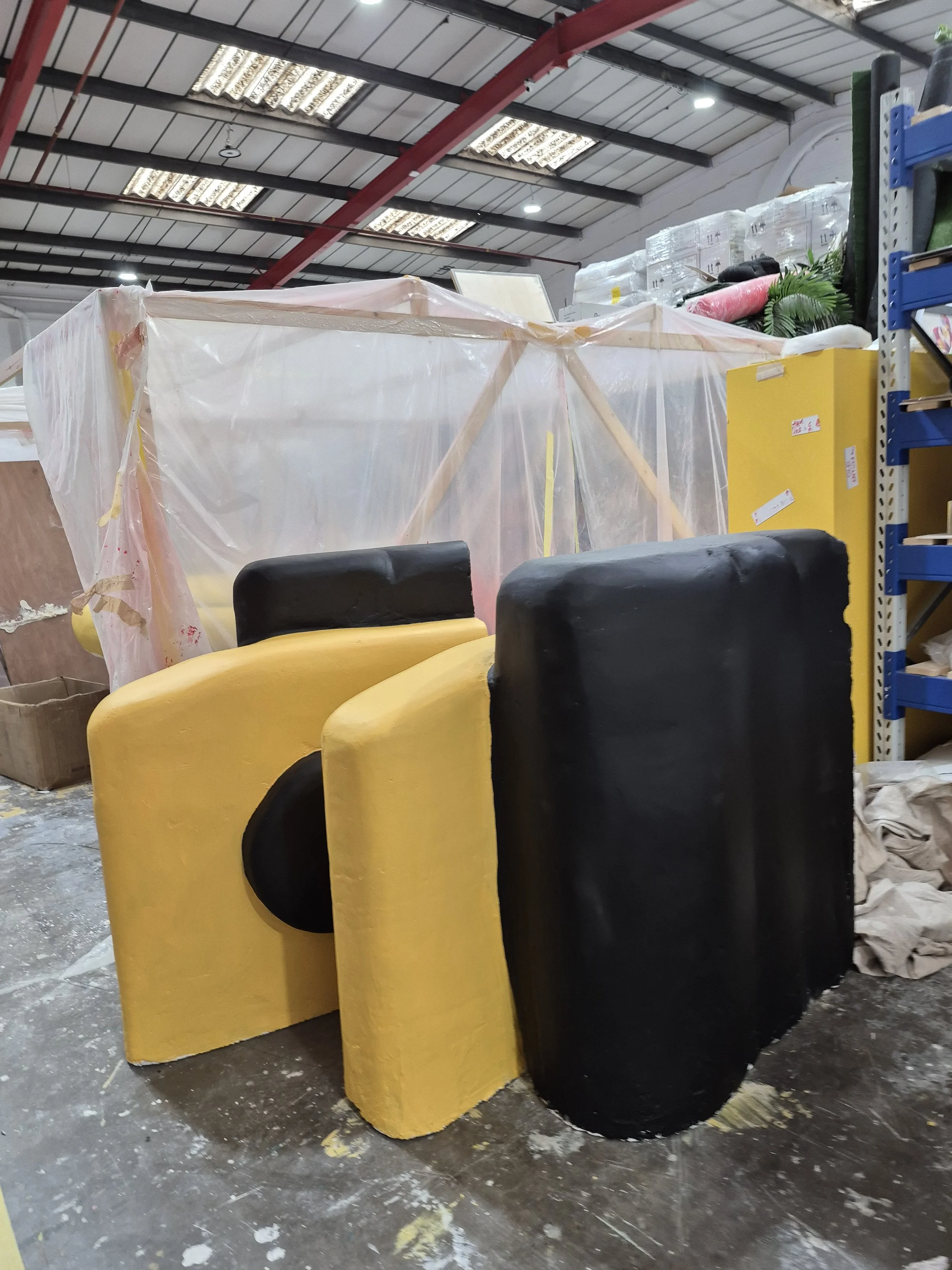 Stacks of foam padding in yellow and black, covered with plastic in a warehouse with shelves and boxes around.