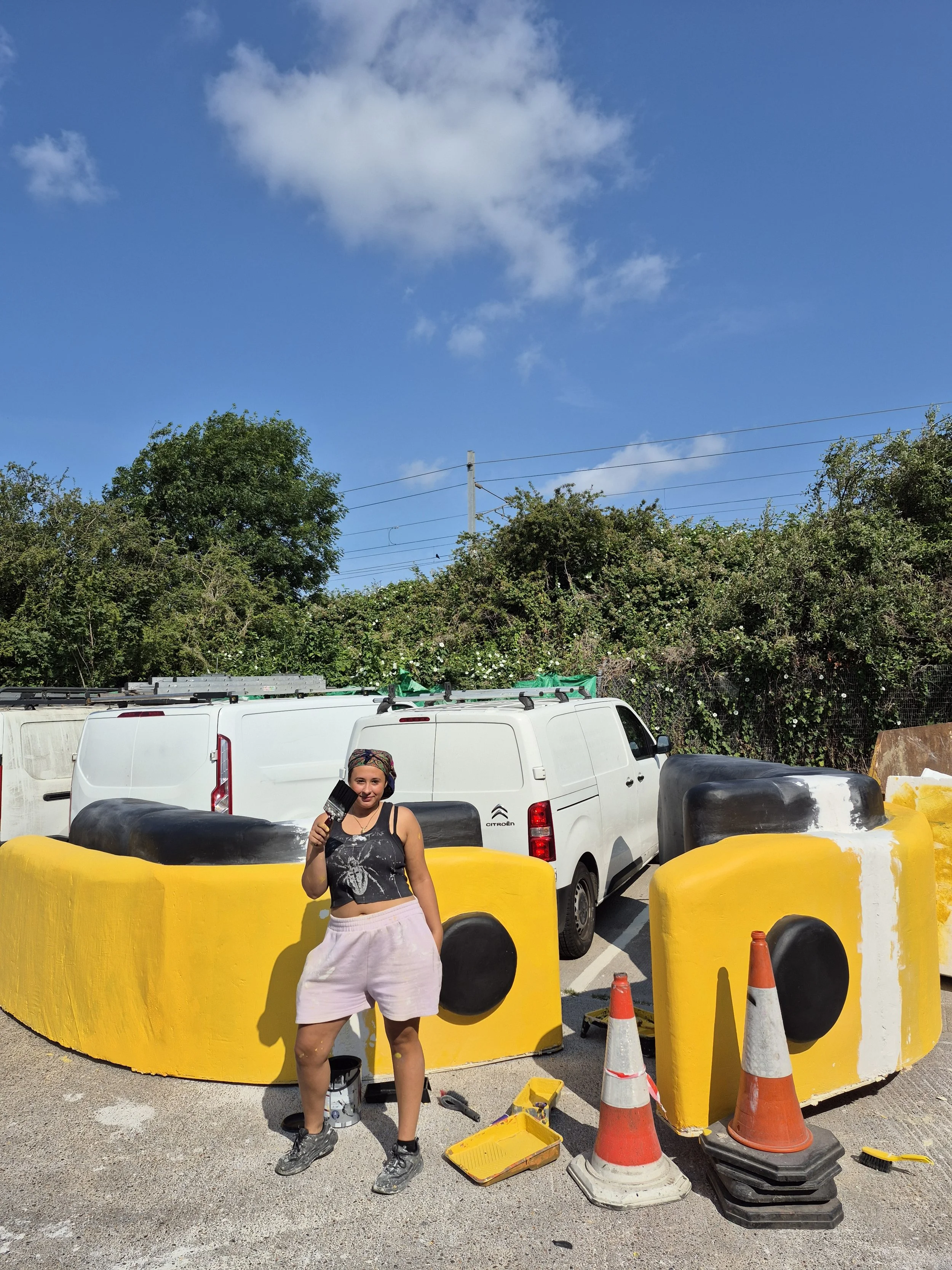 A woman in a black tank top and white shorts holding a paintbrush stands in front of large yellow bumper barriers, with construction tools and traffic cones around her. Behind her, there are white vans, a green hedge, and a blue sky with some clouds.