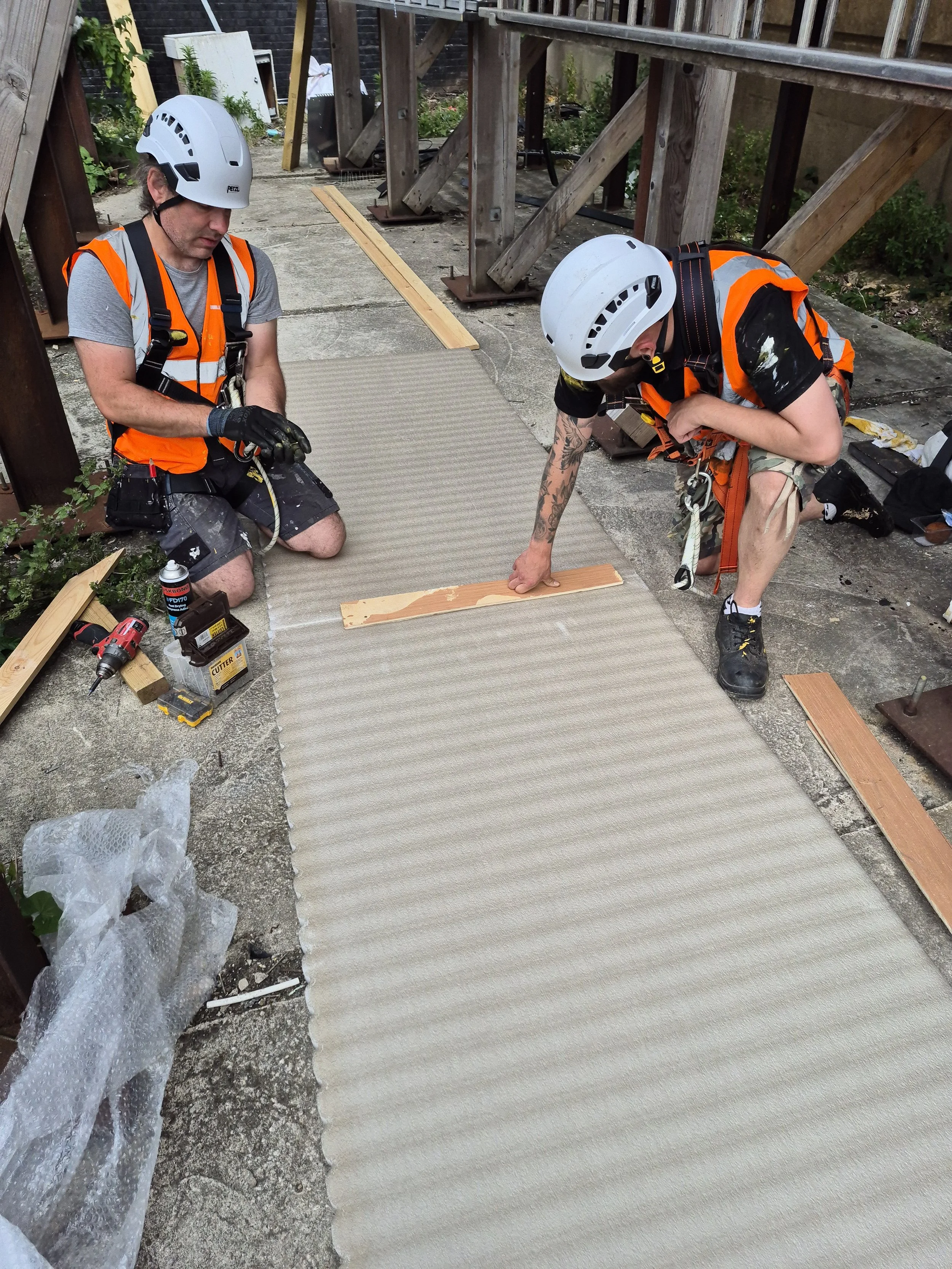 Two construction workers wearing white safety helmets and orange safety vests kneel on a concrete sidewalk, working on a light-colored carpet or mat. One worker is holding a tool, and various tools and materials are scattered around. Wooden planks an
