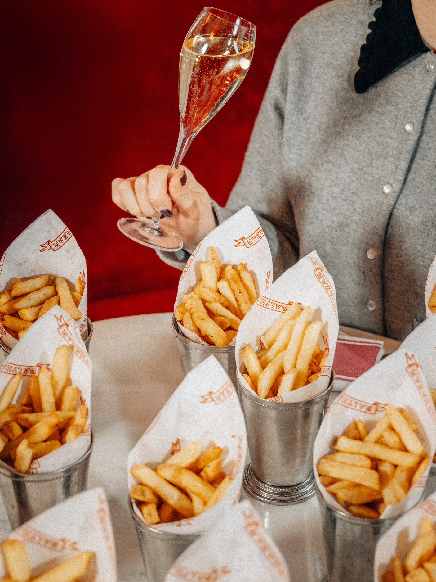 A woman holding a glass of champagne surrounded by several buckets of French fries at a restaurant.