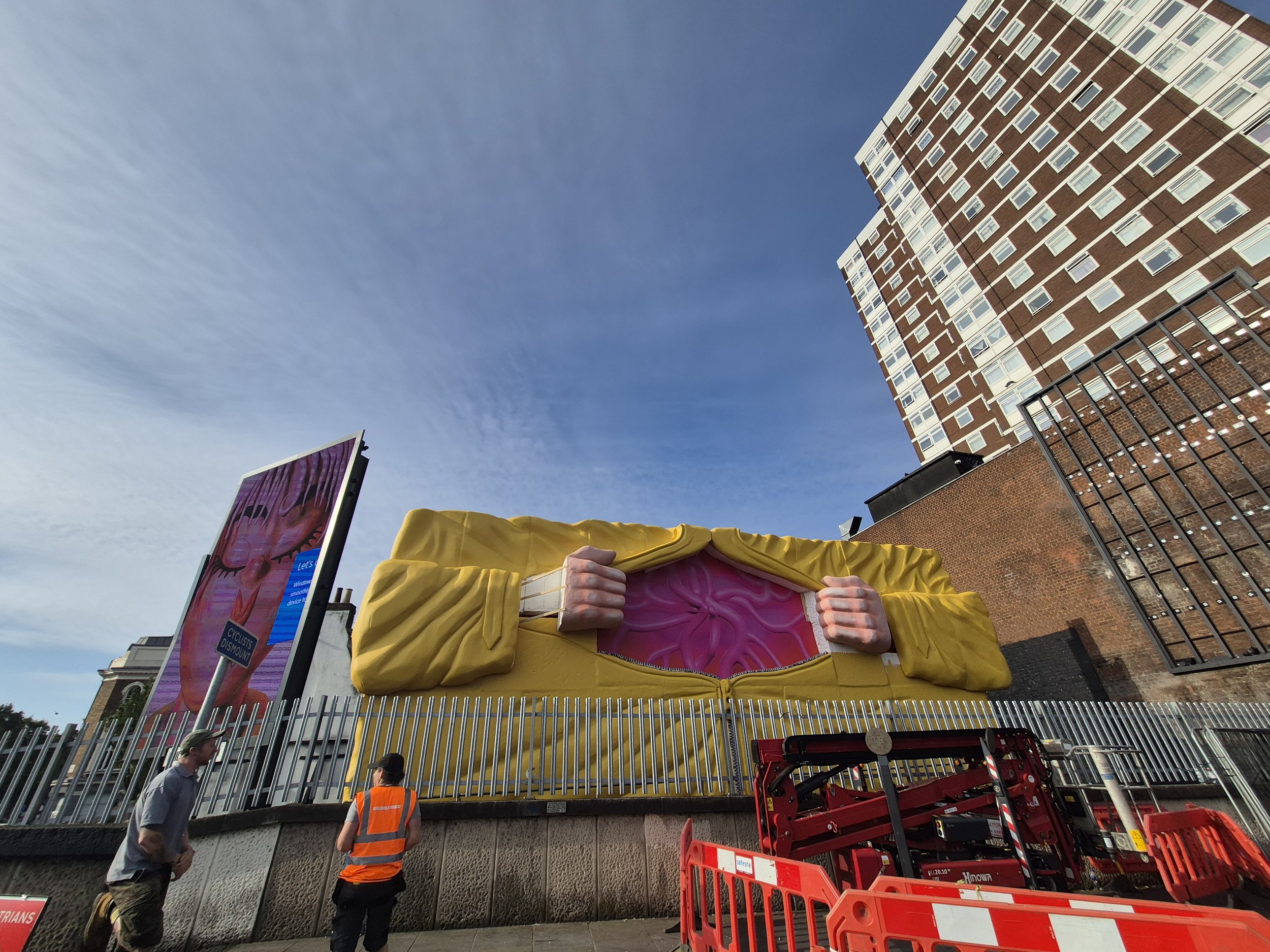 People standing outdoors near a large yellow and pink sculpture resembling a human head with hands, against a backdrop of apartment buildings and a blue sky with some clouds.