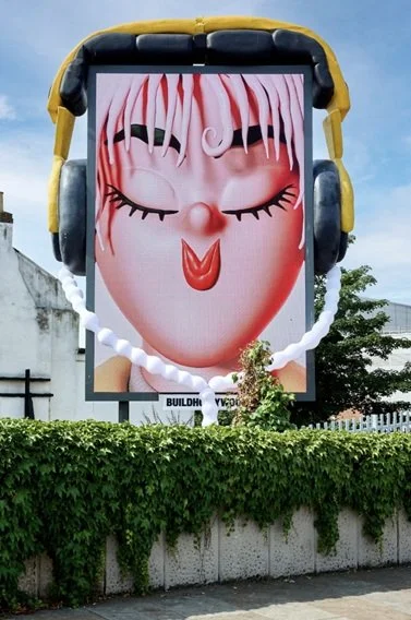Billboard advertising a cartoon face with closed eyes, pink hair, and red lipstick, wearing black headphones, surrounded by a white spiral and greenery.