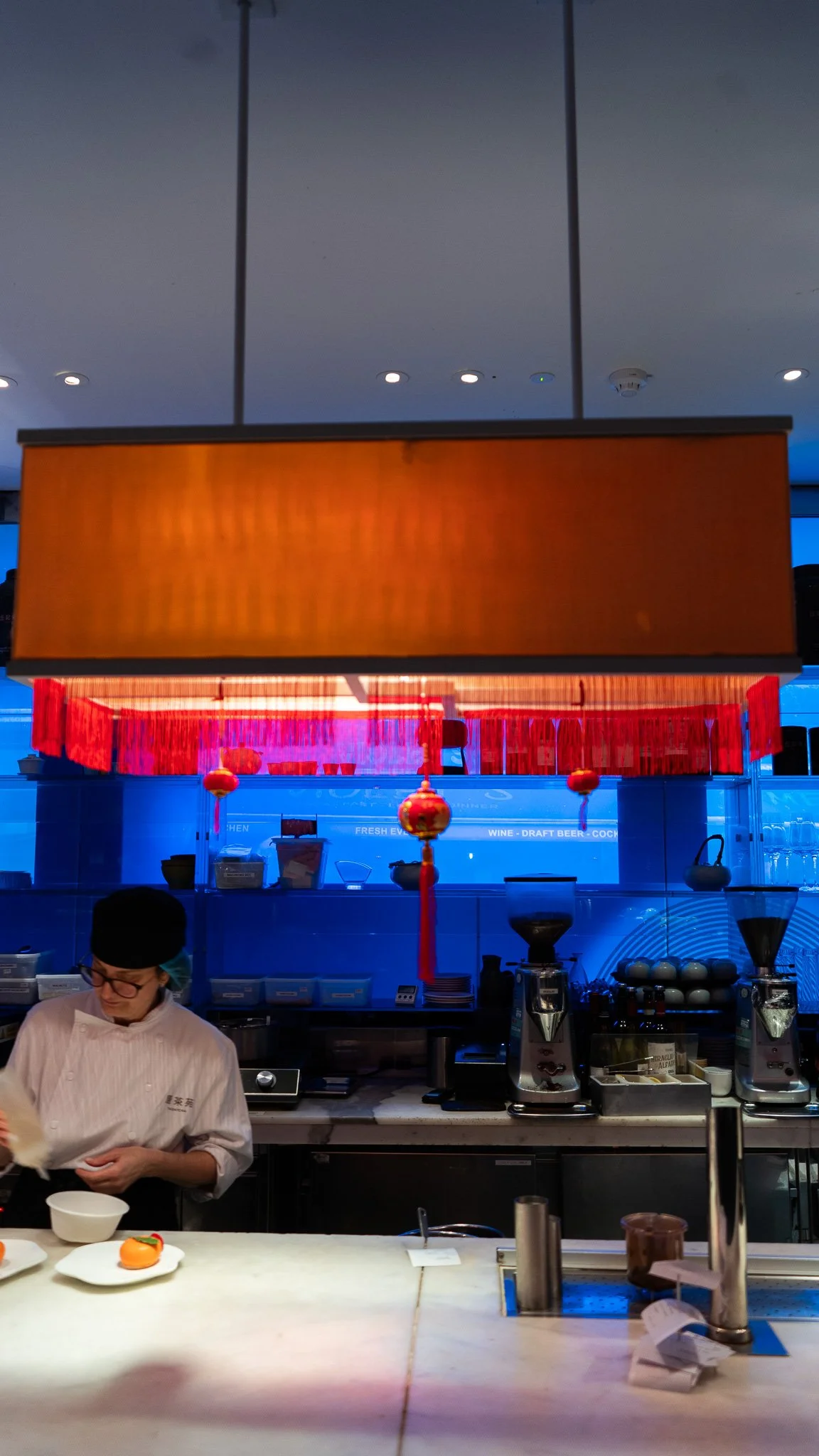 A chef wearing glasses and a white uniform working in a restaurant kitchen with a marble counter and prepared food, with a colorful Chinese lantern hanging from the ceiling above.