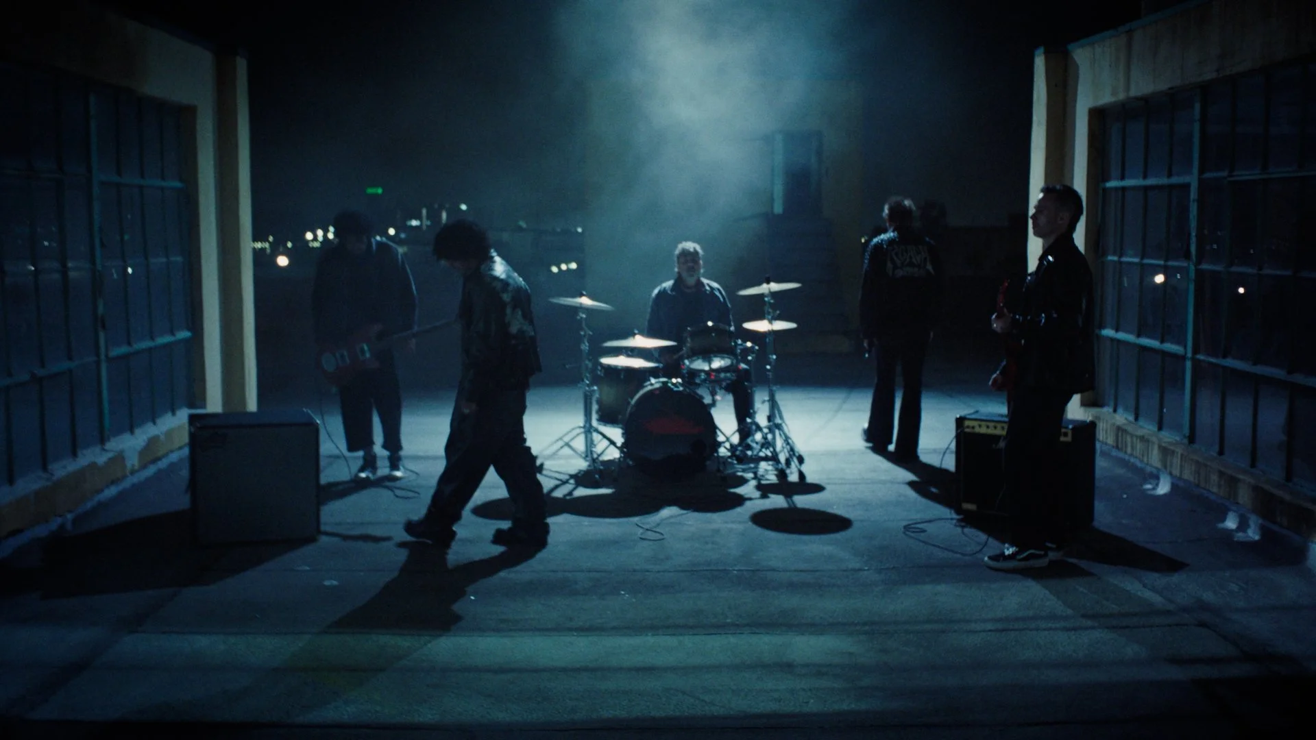 A band of five members performing on a rooftop at night, with the city lights in the background. The scene is dimly lit with a blue hue.