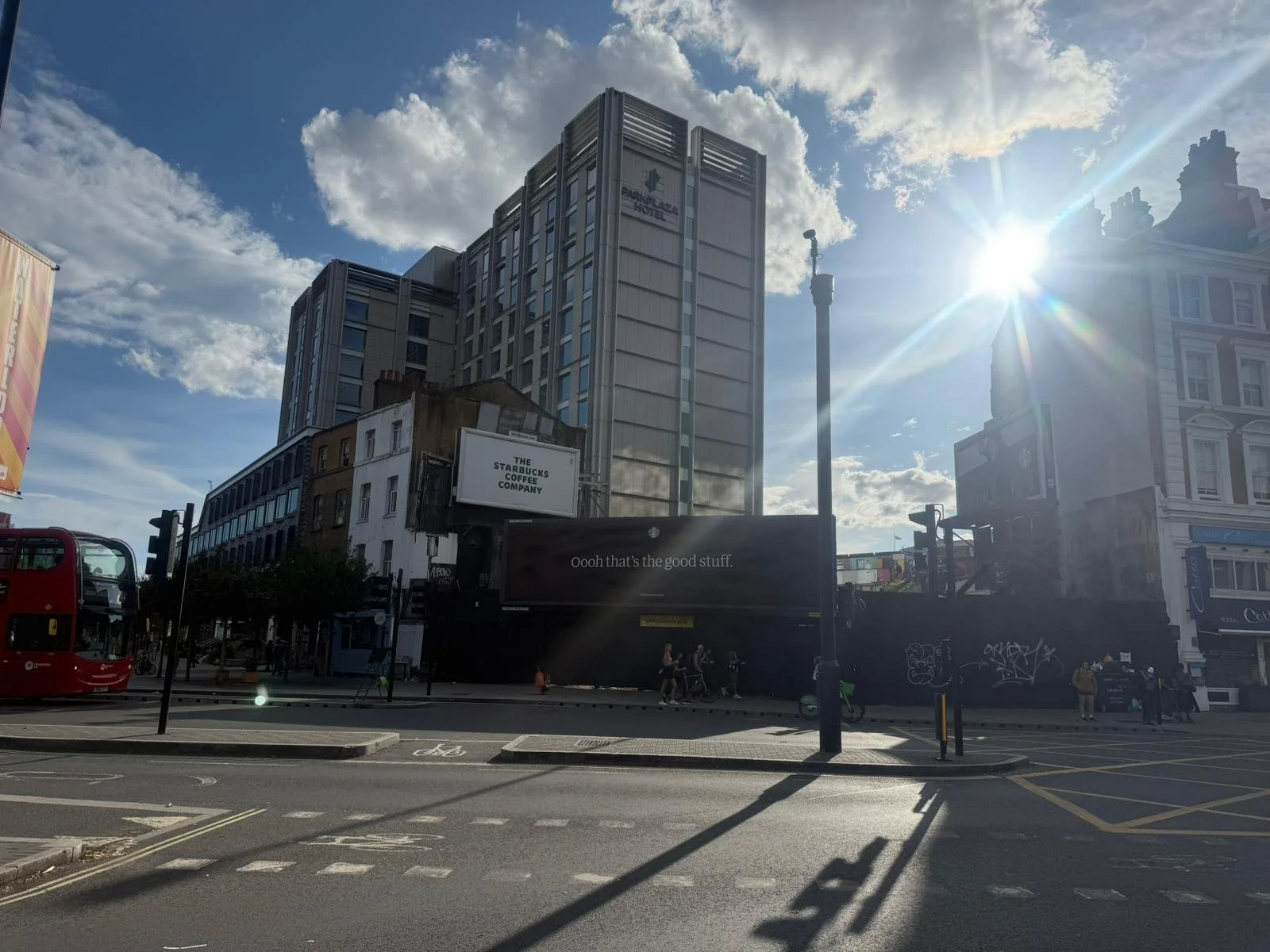 Urban street scene with a modern hotel building, a Starbucks sign, a red double-decker bus, and pedestrians under a partly cloudy sky with sunlight shining through.