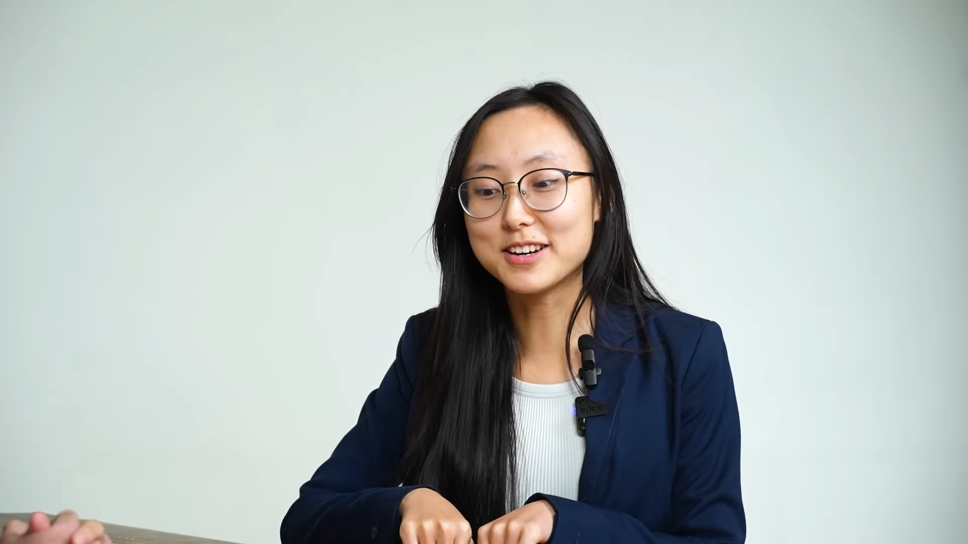 A woman with long black hair, glasses, and a navy blazer is smiling and speaking during an interview or discussion, with a plain light-colored background.
