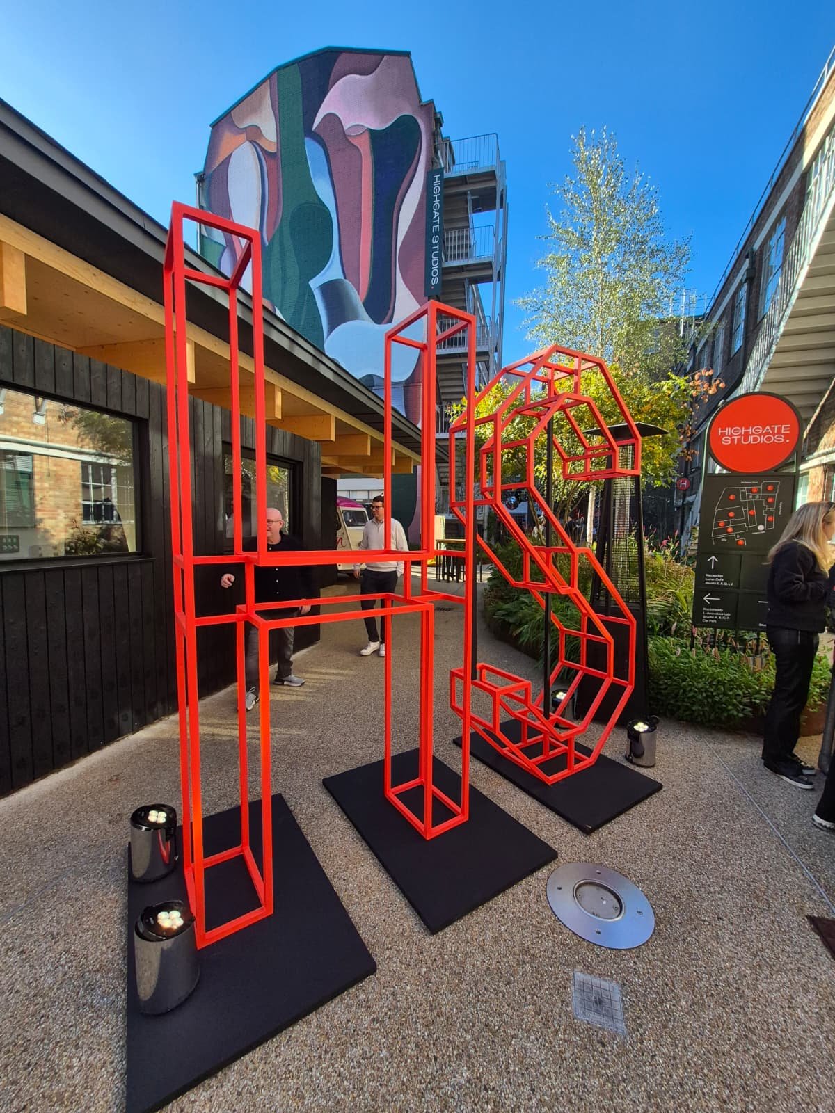 Outdoor art installation featuring red geometric shapes displayed on black platforms with small lights, adjacent to a black building, with people walking nearby and a large colorful mural on a building in the background.