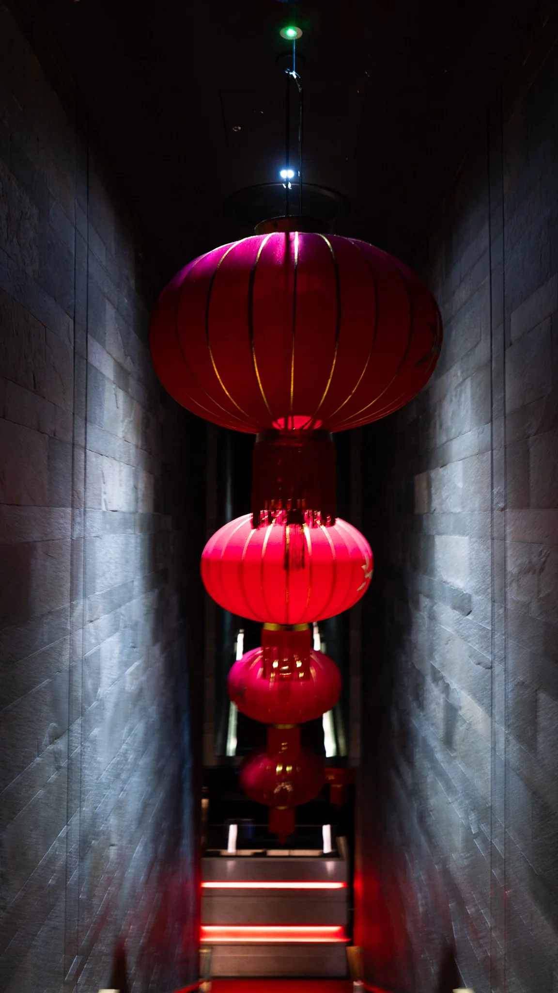 A row of red Chinese lanterns hanging from the ceiling of a narrow corridor with dark stone walls.