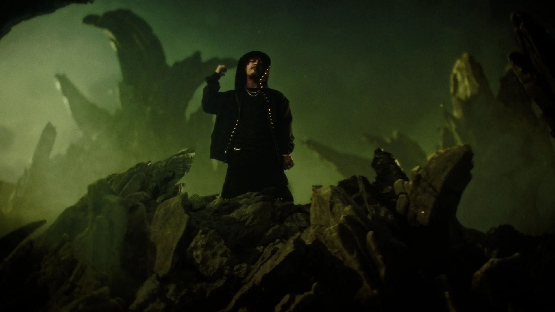 A person wearing a black hoodie and dark clothing standing among large, jagged rocks in a green-hued environment with fog and shadowy structures in the background.
