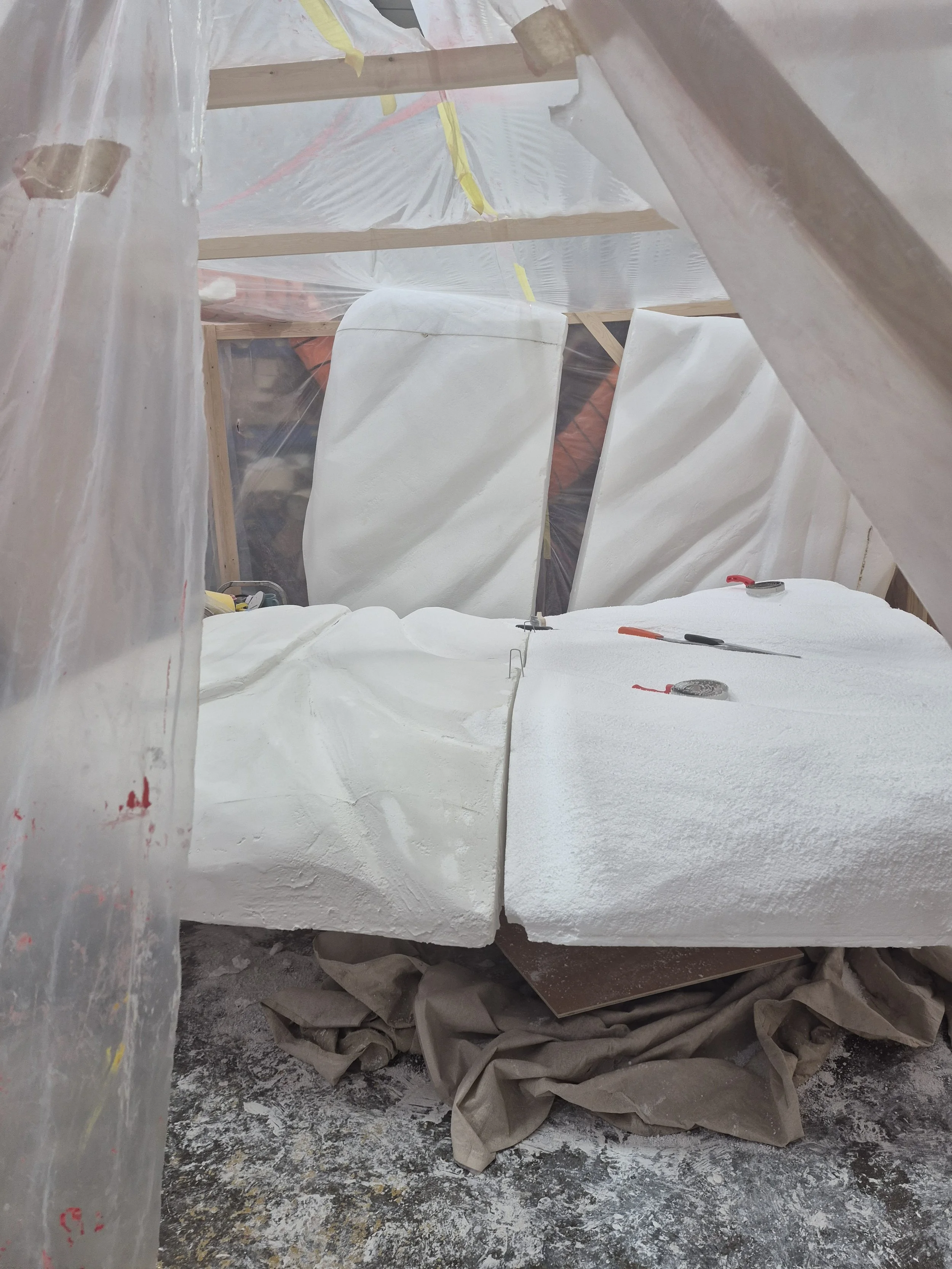 Construction site with white foam insulation sheets, protective plastic coverings, and tools on the floor, surrounded by insulation debris and wrapped in protective plastic sheeting.