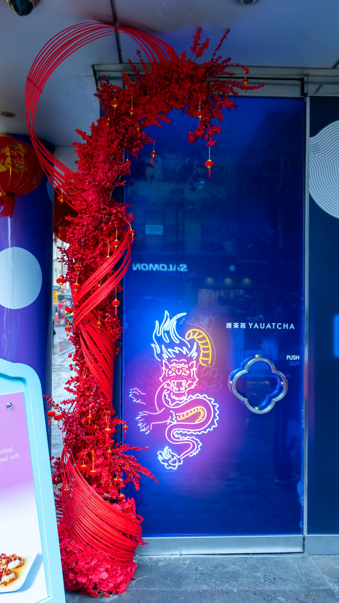 Decorative red floral arrangement with lanterns next to a blue door featuring neon dragon graffiti and a sign for Yuatcha.