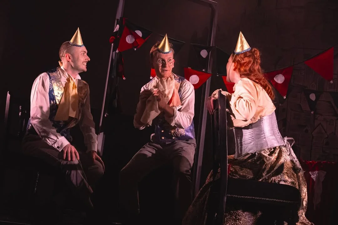 Three performers in costumes with party hats, sitting and standing at a festive event with red and black triangular banners and polka dot decorations.