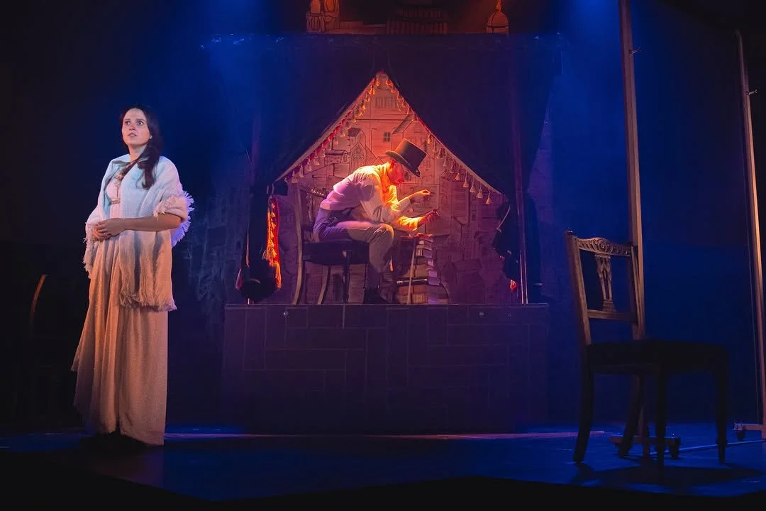 A theatrical scene featuring a woman in a long dress standing to the left and a man in a top hat sitting on a stool on a small stage, with a backdrop resembling a house made of newspaper and string lights.