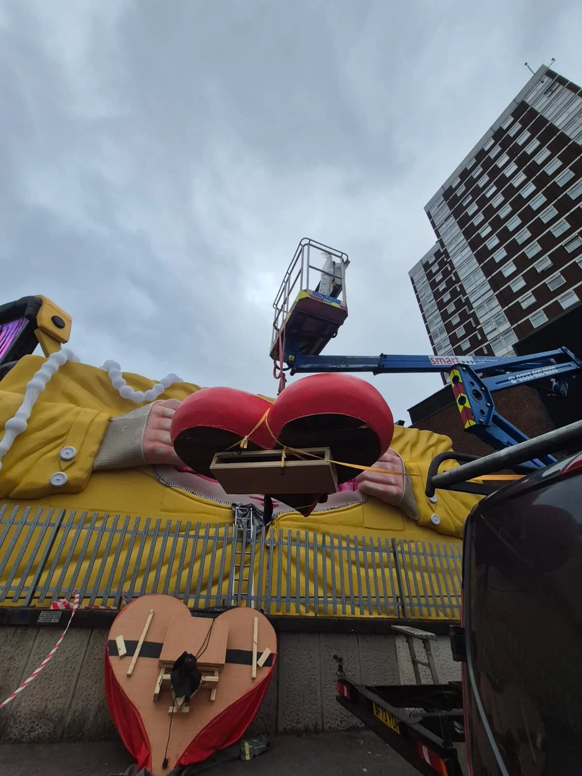 A large yellow float with a heart-shaped cake on top and a giant inflatable figure of a woman in a yellow dress inside. A person is adjusting the float. Overcast sky and tall buildings are in the background.