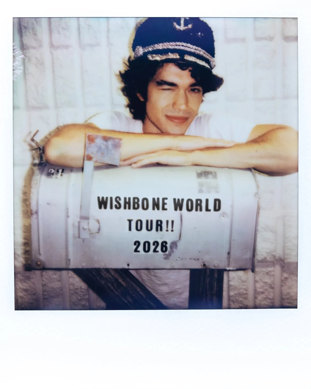 A young man with curly hair wearing a navy sailor hat with an anchor emblem, resting his arms on a metal container that has a sign reading 'Wishbone World Tour!! 2026'. He is winking and smiling, with a light brick wall in the background.