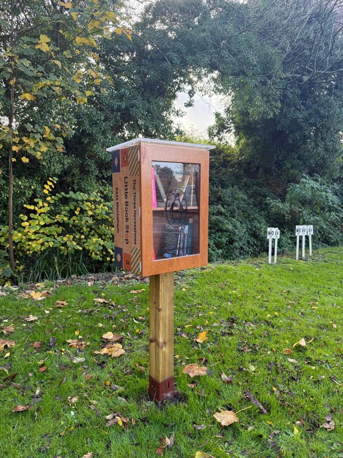 A wooden Little Free Library box labeled "The Three Housings Little Book Stop" is mounted on a wooden post outdoors on a grassy area, with trees and bushes in the background, and three small white signs marked "WOSV" in the distance.