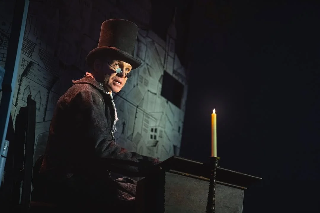 A man dressed in old-fashioned clothing, wearing glasses, a tall top hat, and sitting at a desk with a lit candle, in a dimly lit room with historical drawings on the wall.