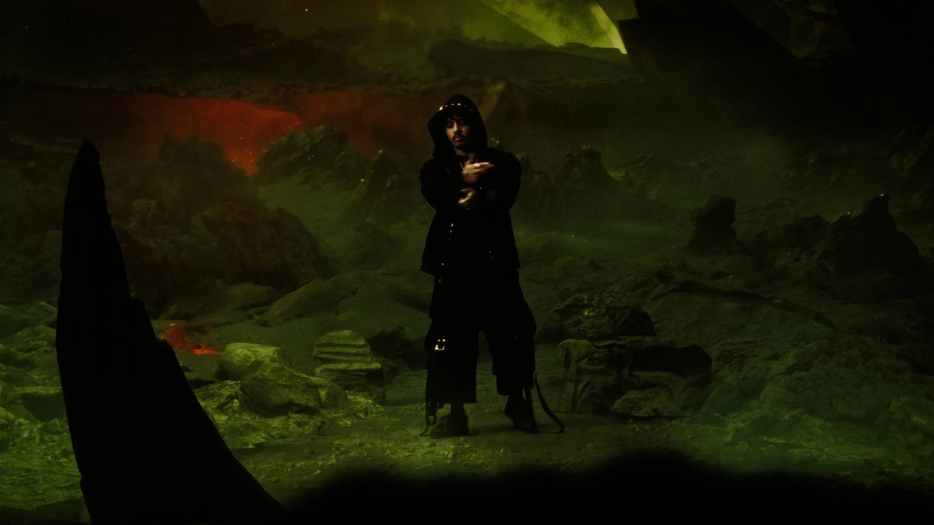 Person standing in a dark, volcanic landscape with glowing lava and greenish hue, wearing black clothing and a hood, with rocky terrain in the background.