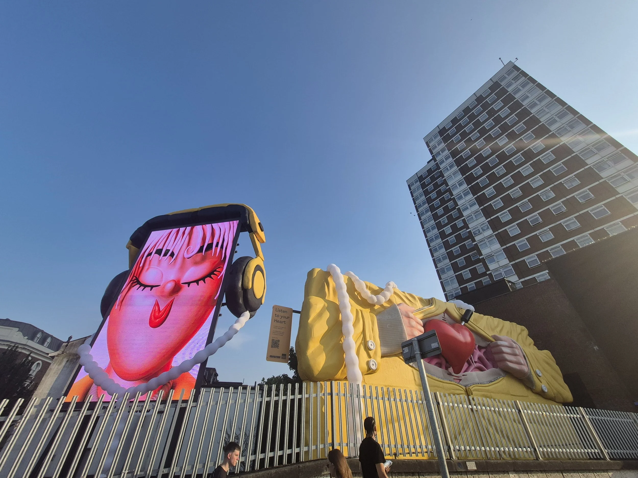 Public art installation featuring a large pink face with closed eyes, set against a blue sky with a modern high-rise building in the background. The sculpture includes yellow and white elements resembling hair and a headpiece, with a small sign readi