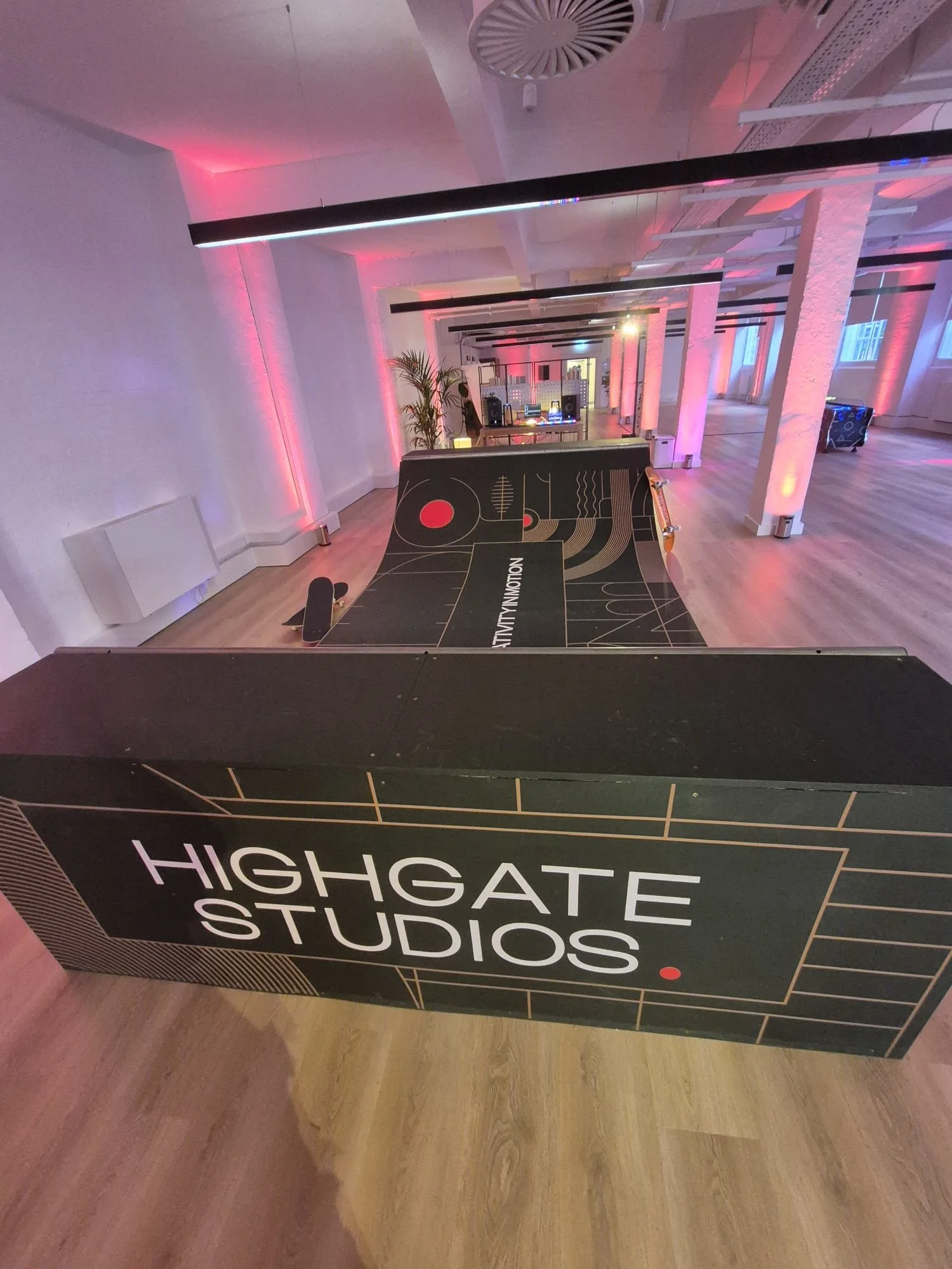A large black sign with white and red text that reads 'Highgate Studios' on a wooden floor inside a studio with white walls, pink and purple lighting, and a DJ setup in the background.