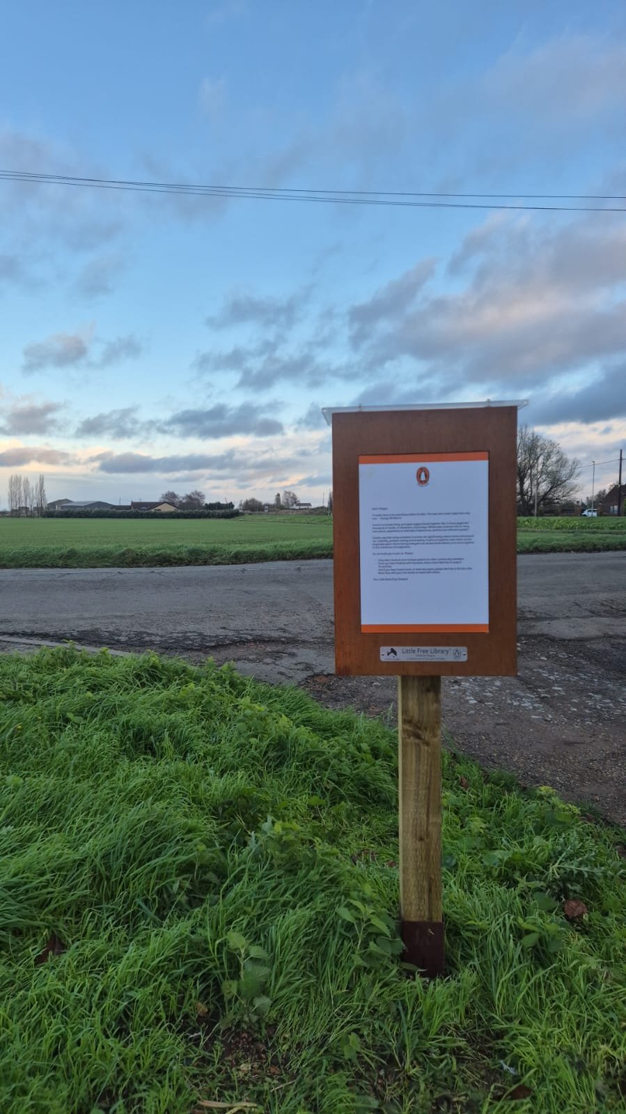 A wooden signboard with a letter on top and a notice below, situated on a grassy area near a paved road in a rural setting with open fields, trees, and a partly cloudy sky.