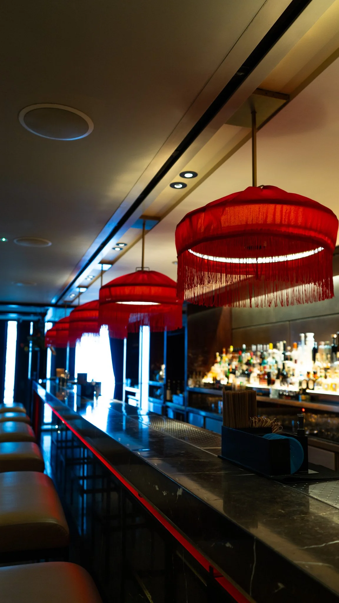 Interior of a bar with a row of red fringed pendant lights hanging above the bar counter, bottles of liquor in the background, and bar stools along the counter.