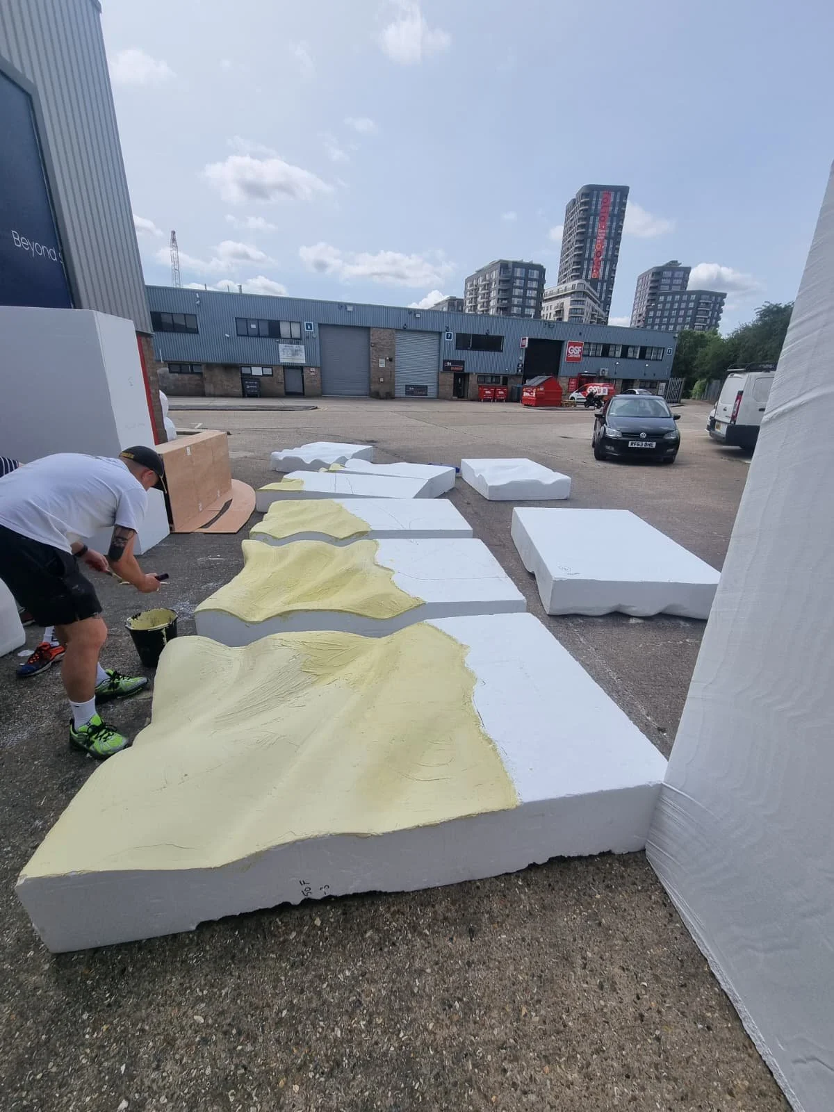 Person painting large foam or sculpture pieces outdoors, with a cityscape background including tall buildings and cloudy sky.