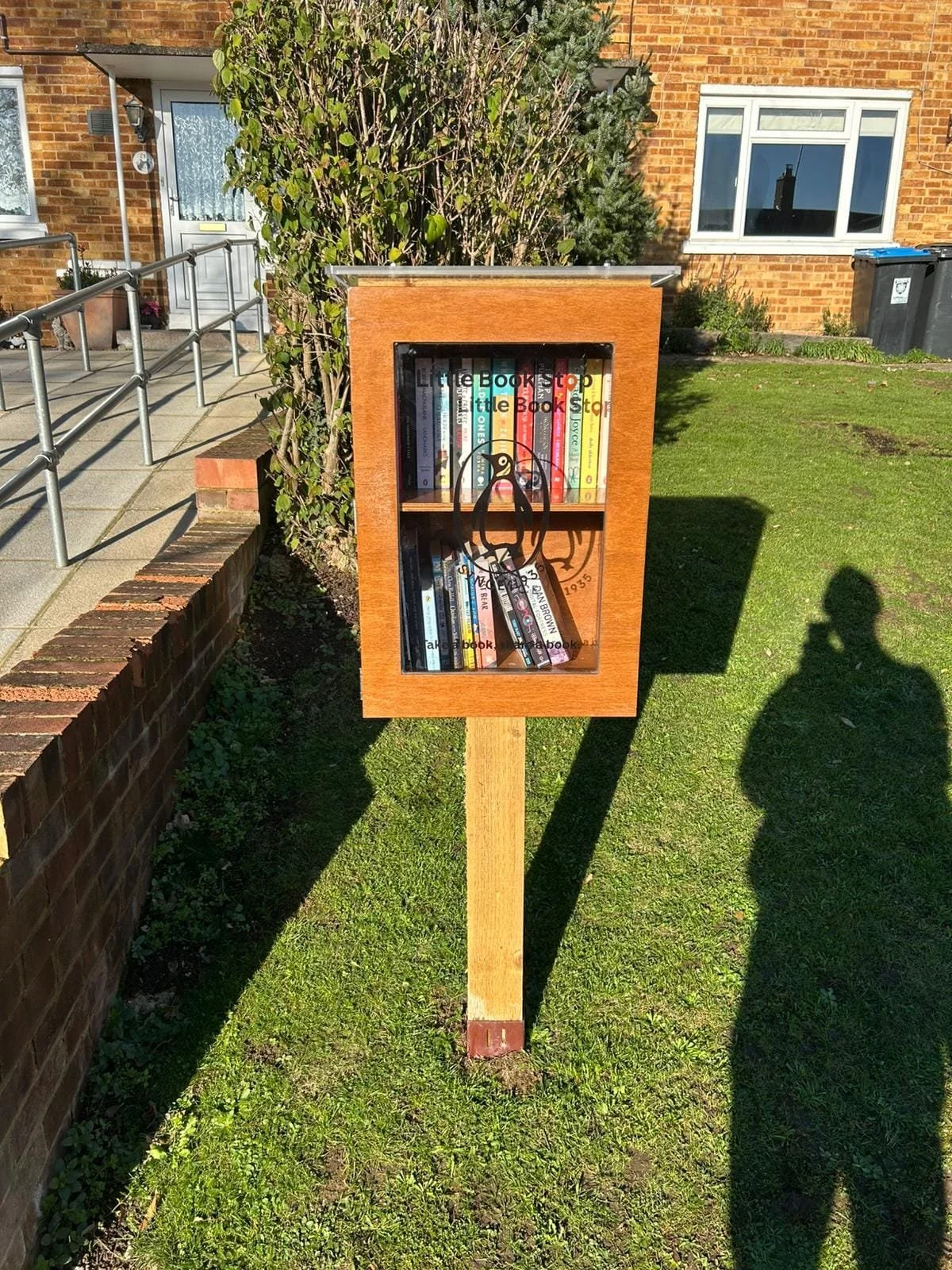 Little Free Library mounted on a post in a residential yard. The library has a glass front, filled with books, and a bird illustration on the door. The yard is grassy with a brick wall, a house with brick exterior, and a bush in the background.