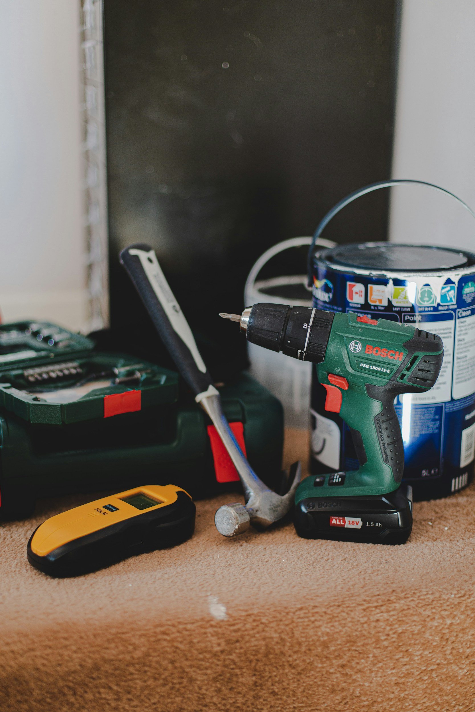 A cluttered workspace with a Bosch cordless drill, a yellow laser level, a cordless screwdriver, a paint can, a pen, and other tools on a carpeted surface.