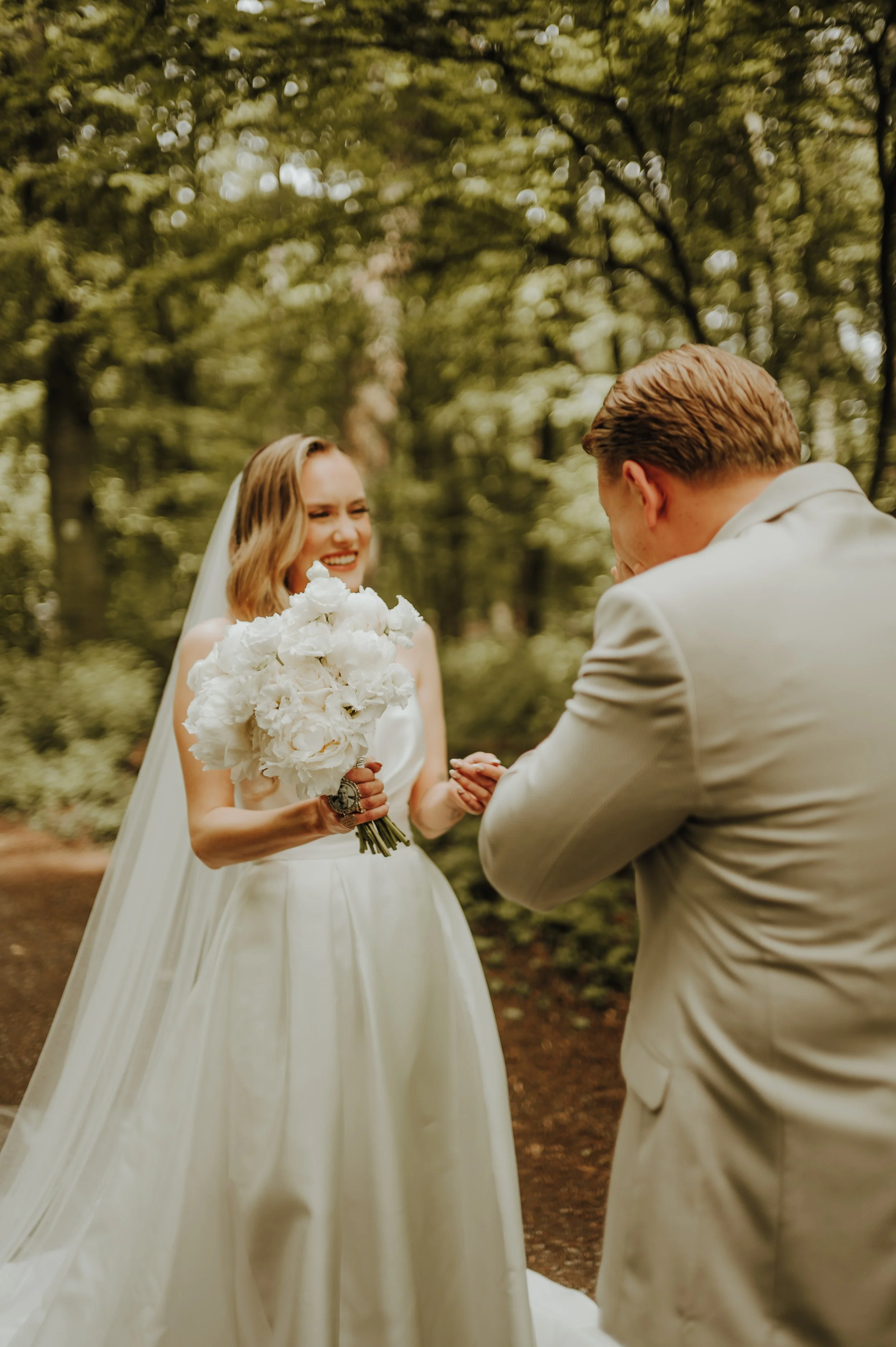 Ein Brautpaar bei einer standesamtlichen Hochzeit im Freien, der Fokus liegt auf der Braut, die ein weißes Kleid und einen Brautstrauß in der Hand hält, während der Bräutigam vor ihr steht, beide sind in einem Wald im Sommer. Der Moment scheint emotional und schön.