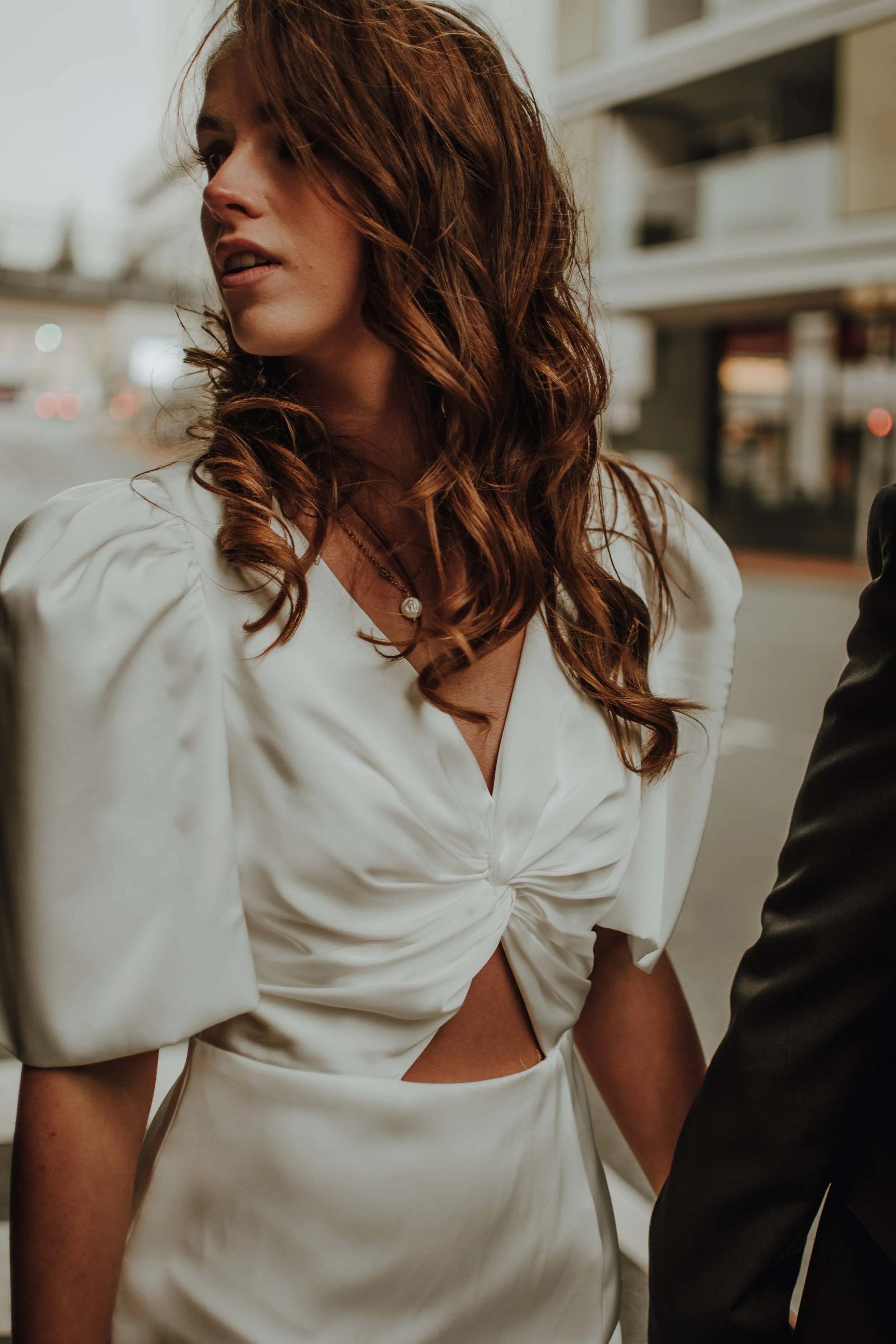 Nahaufnahme eines Frauen-Kleid in Weiß mit tiefem Ausschnitt und Knopfdetail, rote Locken, sichtbares Halskettens, während sie nach links schaut, im Hintergrund eine urbanen Umgebung.