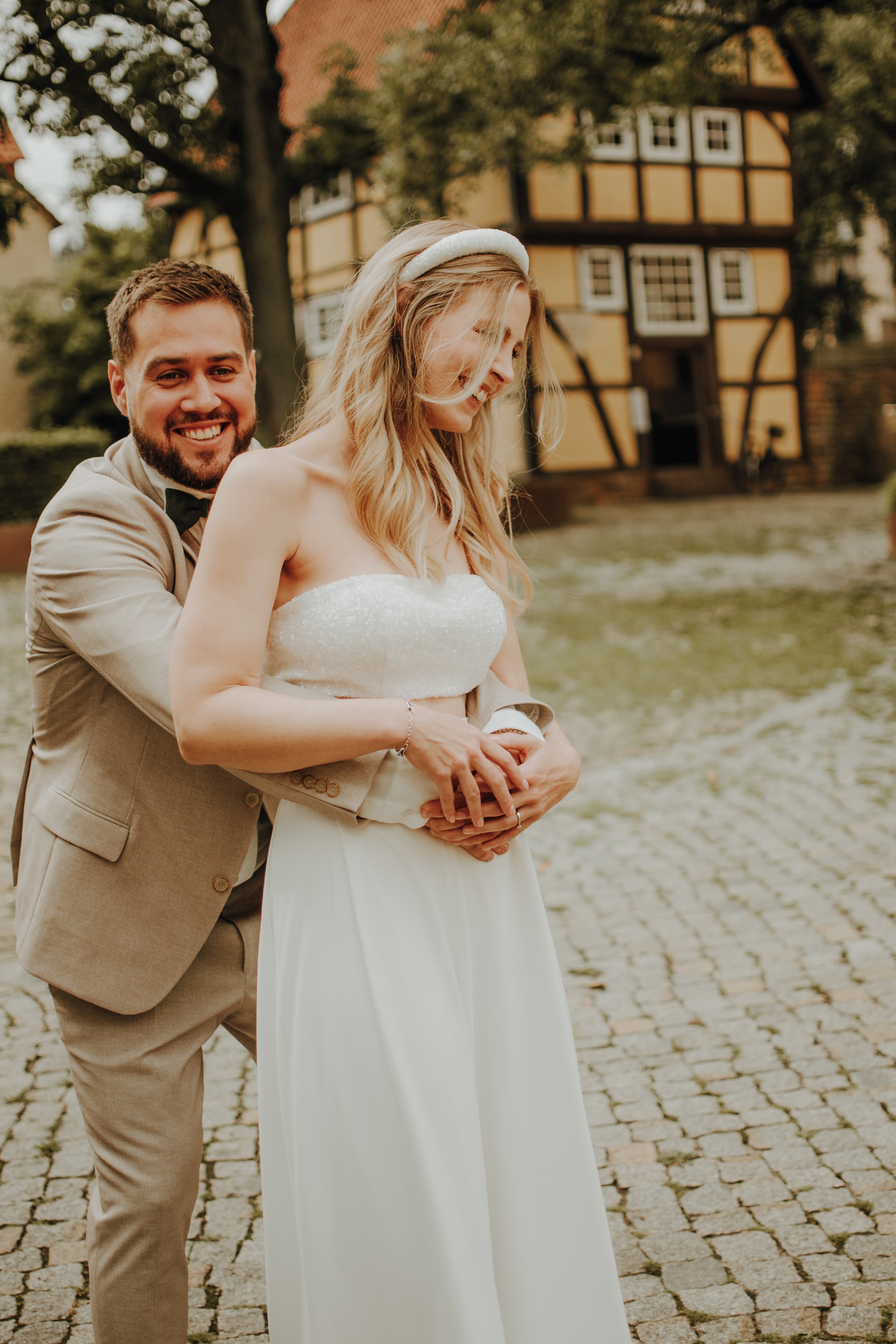 Ein Brautpaar bei einer Hochzeit draußen, im Hintergrund ein historisches Fachwerkhaus, die Frau trägt ein weißes Brautkleid, der Mann einen hellen Anzug.