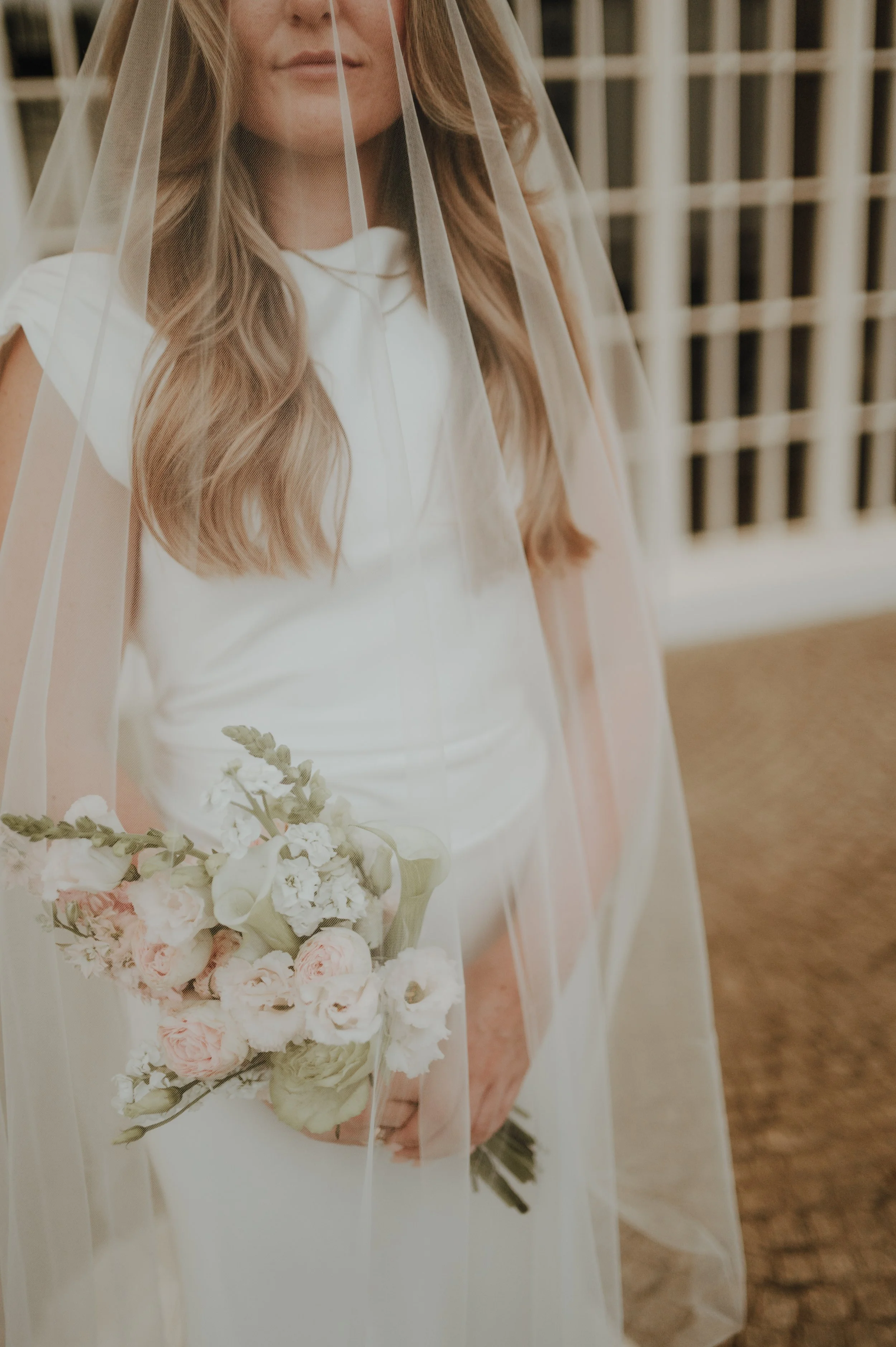 Nahaufnahme einer Frau in Brautkleid mit Schleier, die einen Brautstrauß aus weißen und hellrosa Blumen hält, im Hintergrund eine Wände mit Holzlisen.