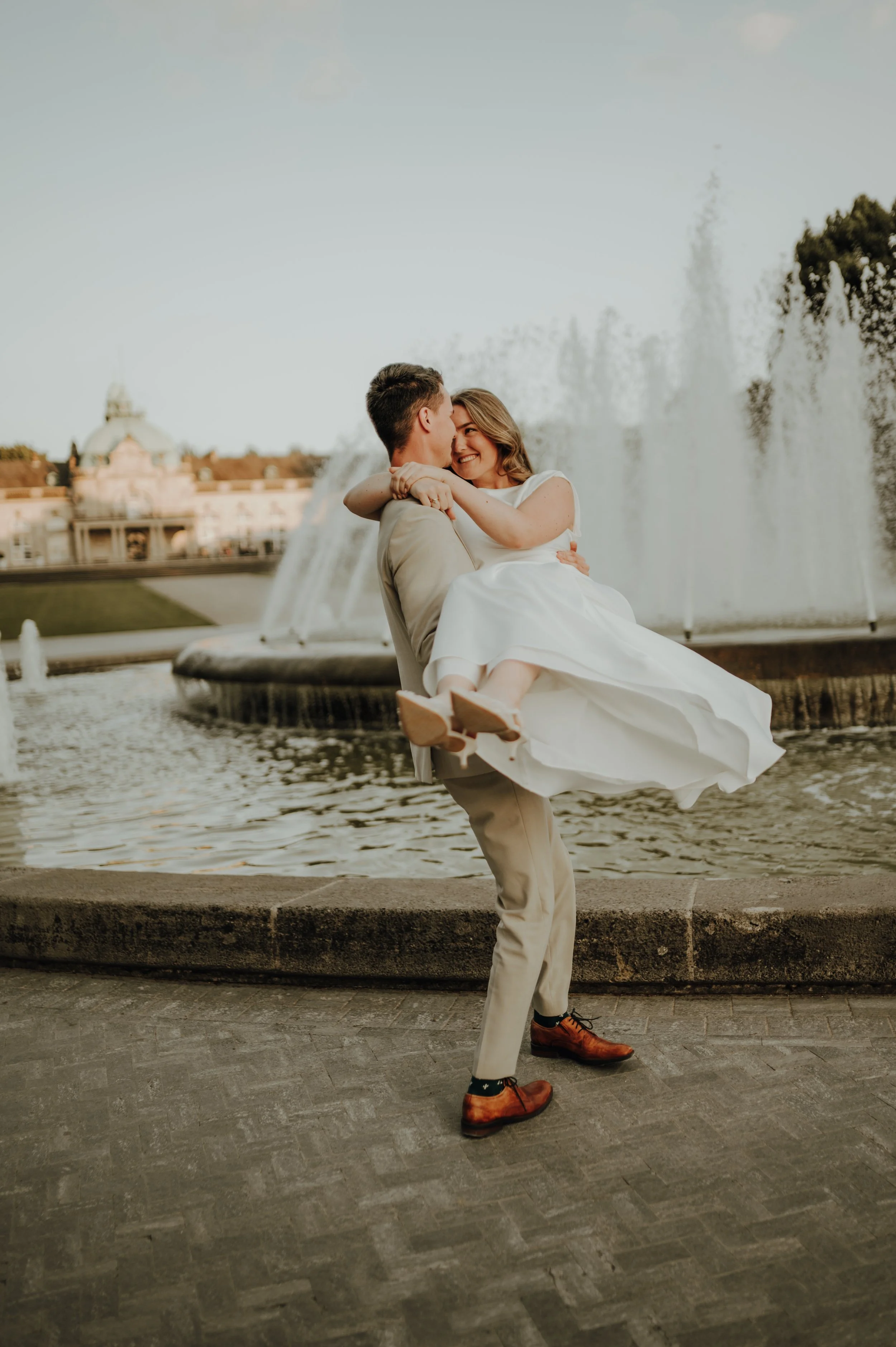 Ein lachendes Paar, wobei der Mann die Frau hochhebt, vor einem Wasserbrunnen im Freien, bei Sonnenuntergang, im Dresscode für eine Hochzeit.