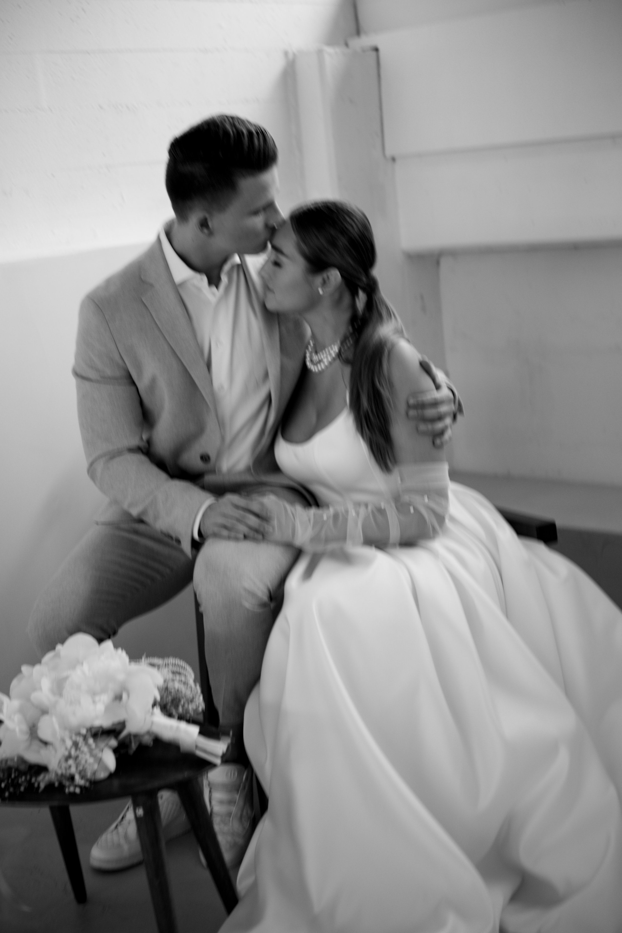 Ein junges Paar bei einer Hochzeit, sie umarmt und küsst sich in einem Raum mit weißen Wänden, Blumen auf einem Tisch, sie trägt ein weißes Brautkleid und er einen hellen Anzug.
