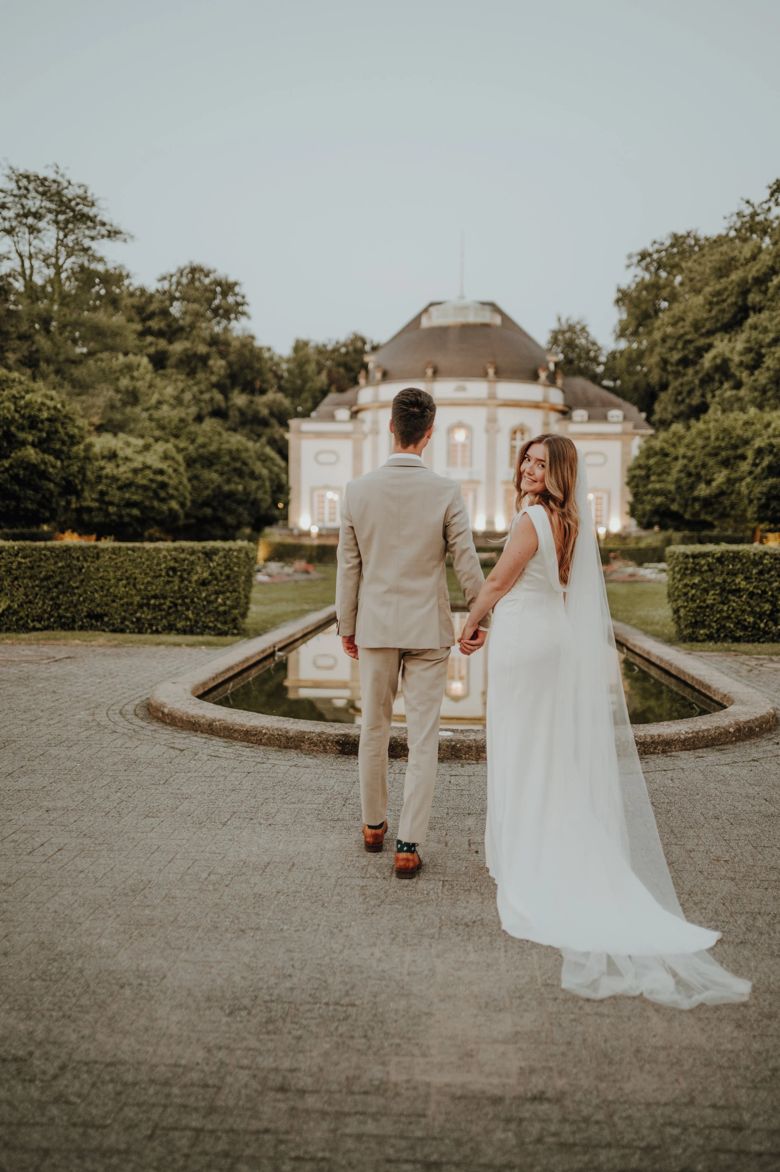 Ein Brautpaar, das sich an einem Abend, vermutlich bei Sonnenuntergang, vor einem historisch aussehenden Gebäude im Park-Umfeld hält und Hand in Hand geht, während die Braut in einem weißen Hochzeitskleid und der Bräutigam in einem hellen Anzug gekleidet sind.