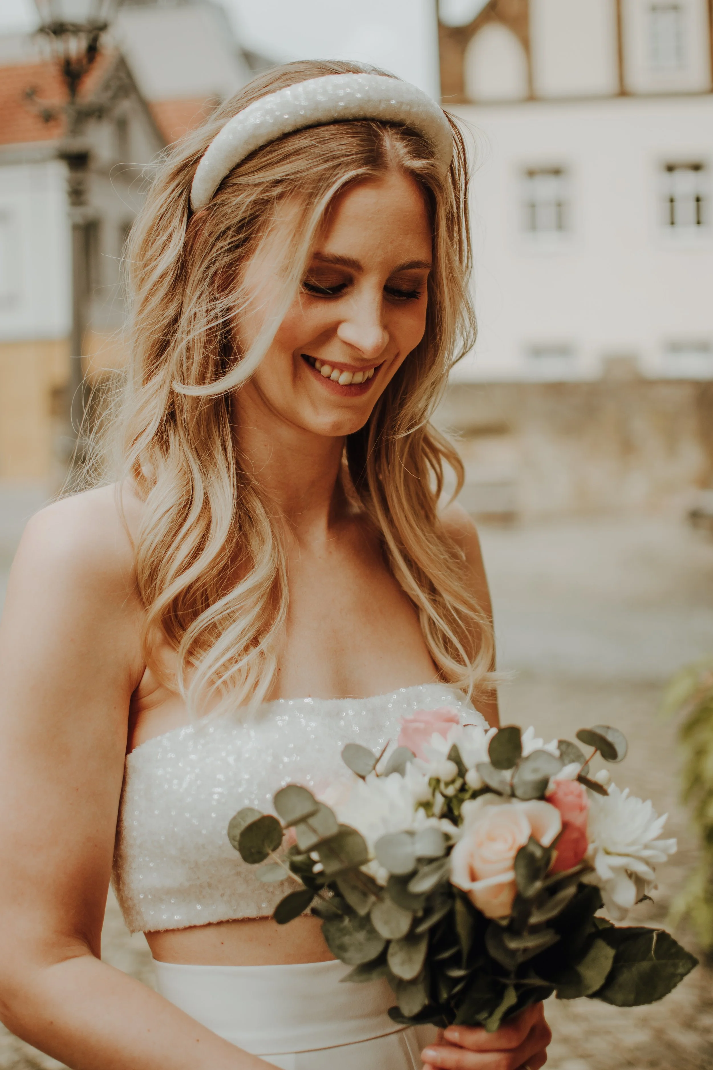 Junge Braut mit Zopf, weißem Haarband und Hochzeitskleid hält einen Blumenstrauß mit Rosen und Eukalyptus, draußen bei sonnigem Wetter.