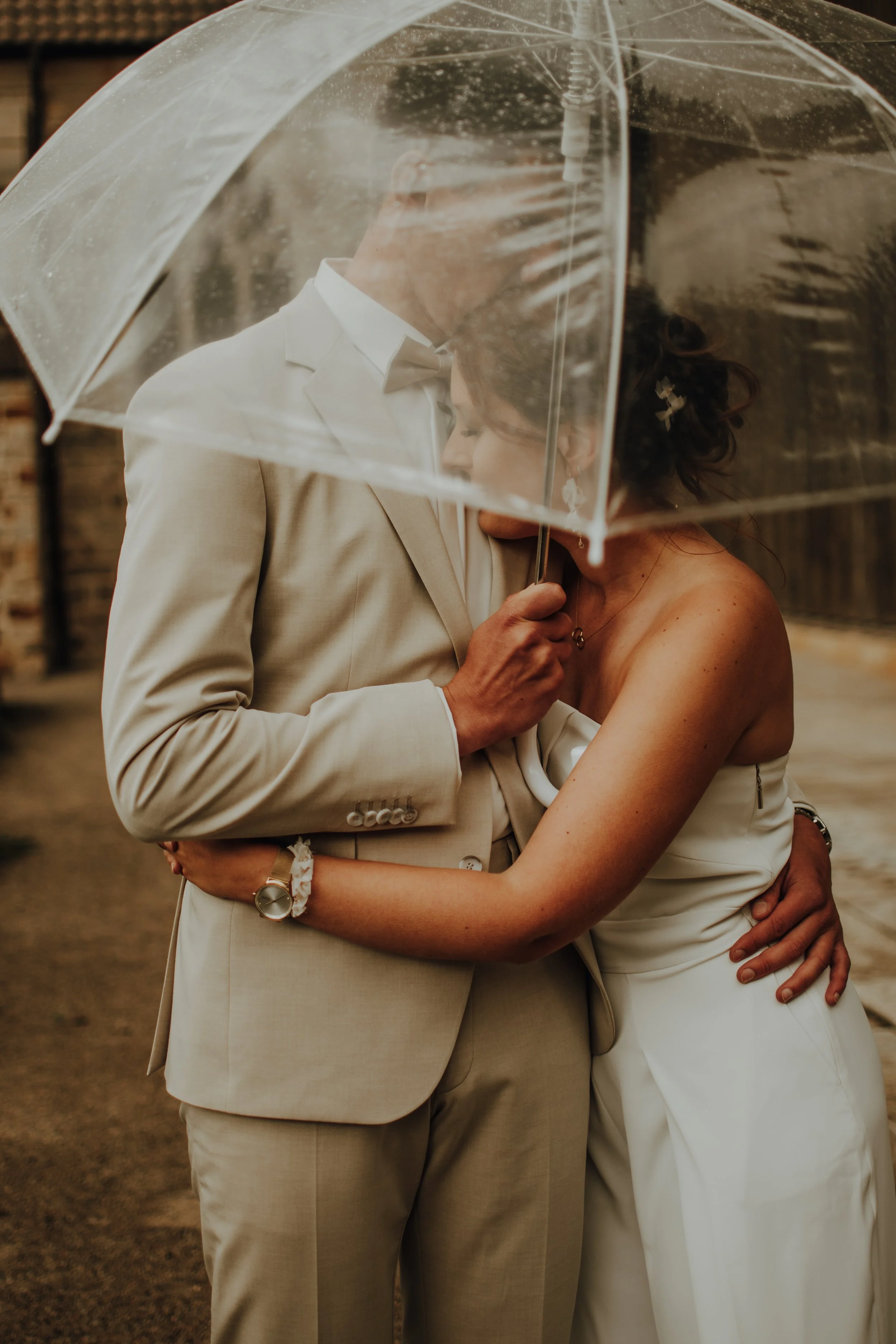 Braut und Bräutigam teilen ein inniges Kuss- und Umarmungsfoto unter einem transparenten Regenschirm bei Regen, im Hintergrund eine Holzwand, bei einer Hochzeitsfeier im Freien.