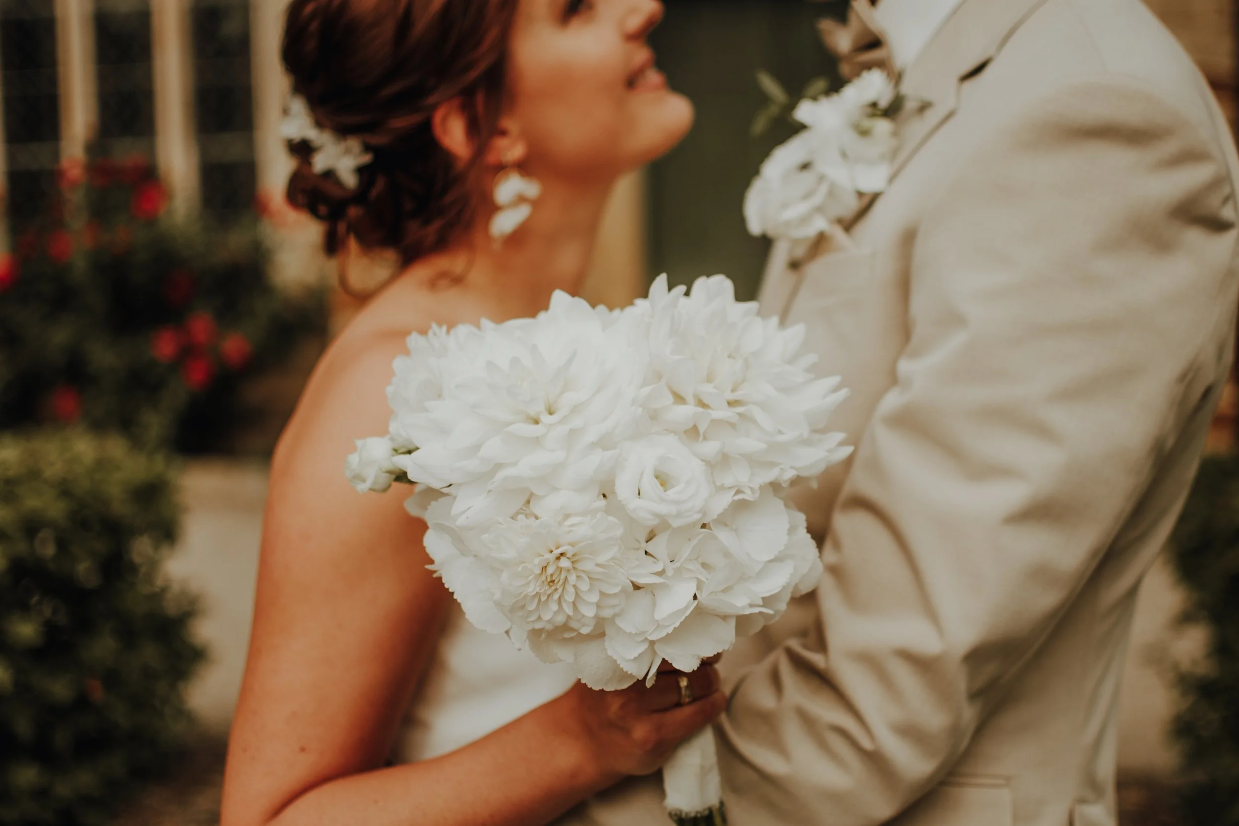 Ein Brautpaar bei einer Hochzeit. Die Braut trägt eine weiße Blume in den Haaren und hält einen großen Strauss weißer Blumen. Das Bild ist unscharf, Fokus auf die Blumen, mit einem Graf Hintergrund.