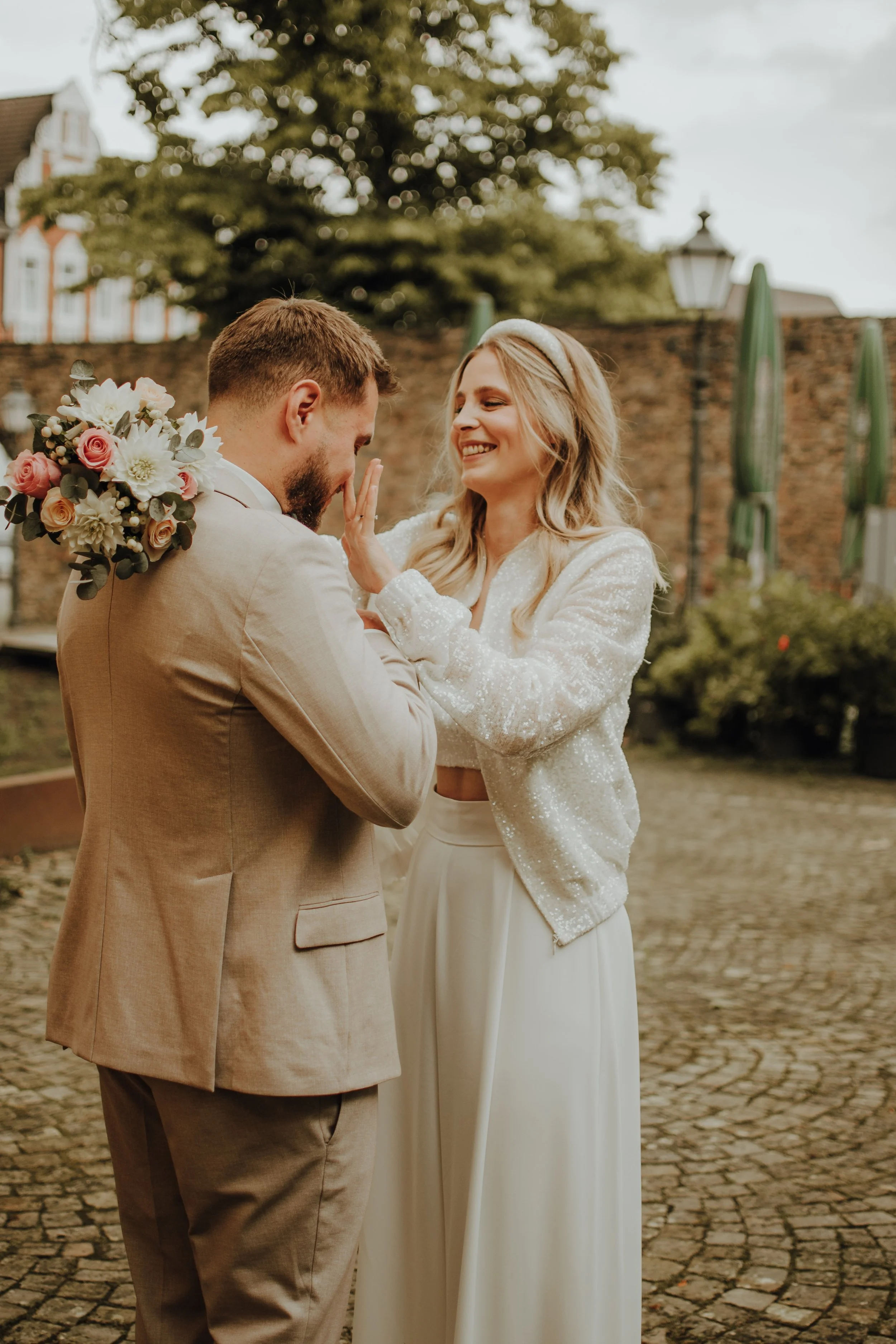 Ein Paar bei einer Hochzeit, das sich liebevoll ansieht und umarmt, im Hintergrund Bäume und ein altes Gebäude.