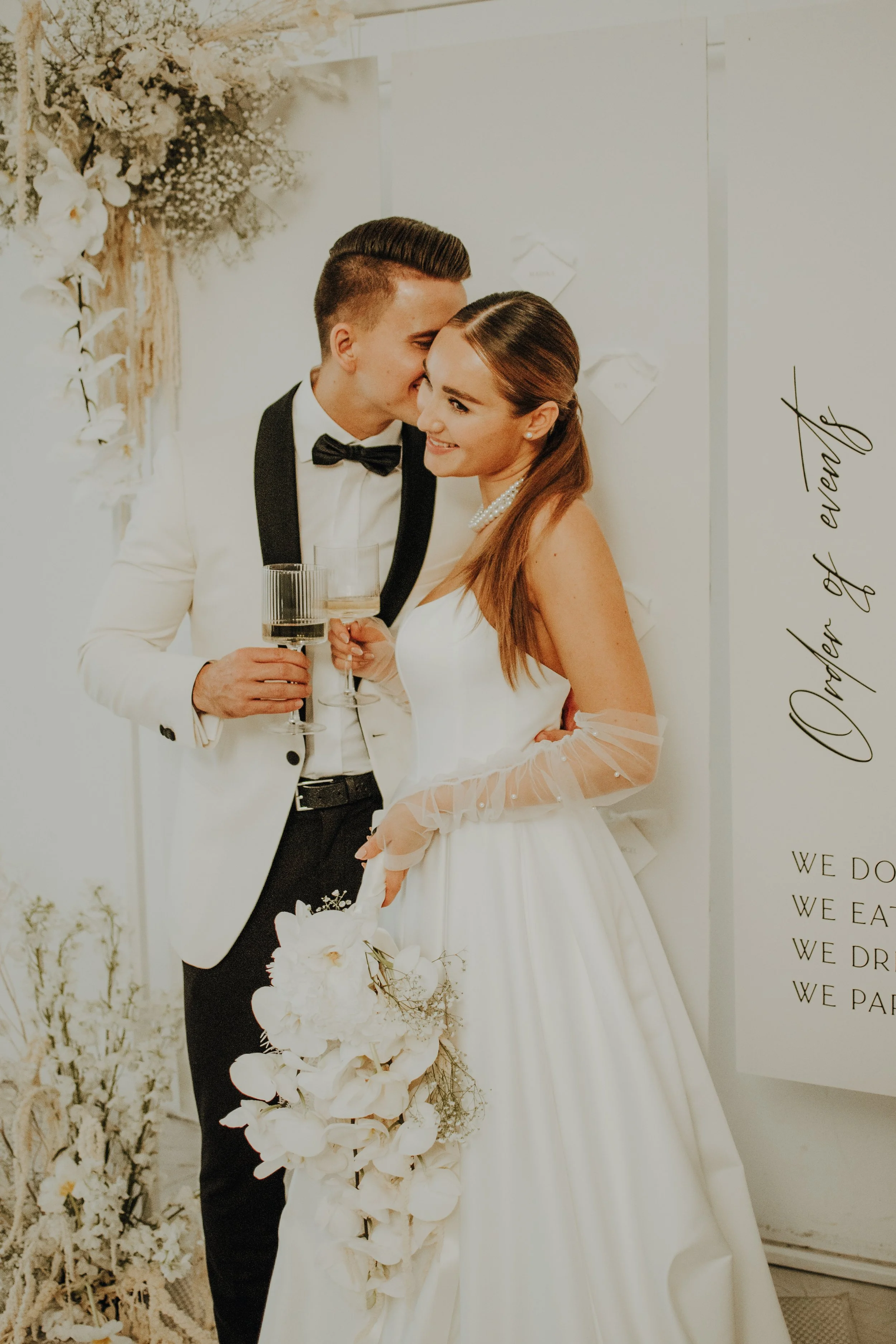 Ein frisch verheiratetes Paar bei ihrer Hochzeitsfeier, der Bräutigam küsst die Braut auf die Stirn, beide halten Gläser mit Getränken, im Hintergrund weiße Dekorationen und Blumen, sehr elegantes Hochzeitsoutfit, die Braut trägt Perlenkette und ein weißes Brautkleid, die Stimmung ist fröhlich und liebevoll.