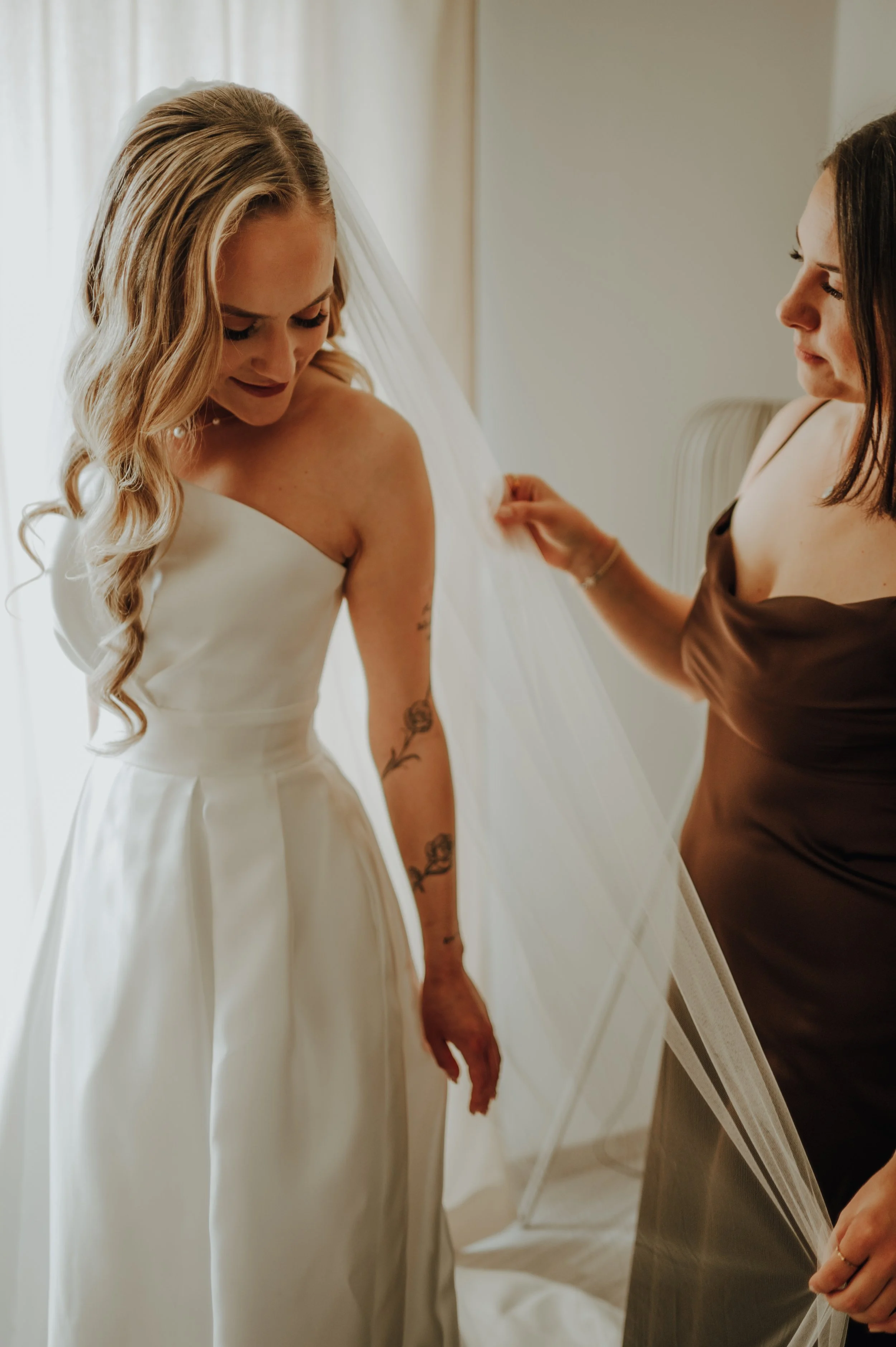 Zwei Frauen, eine in einem weißen Kleid mit langen Locken, die andere in einem braunen Kleid, die eine Frau beim Anziehen eines Schleiers für eine Hochzeit hilft.