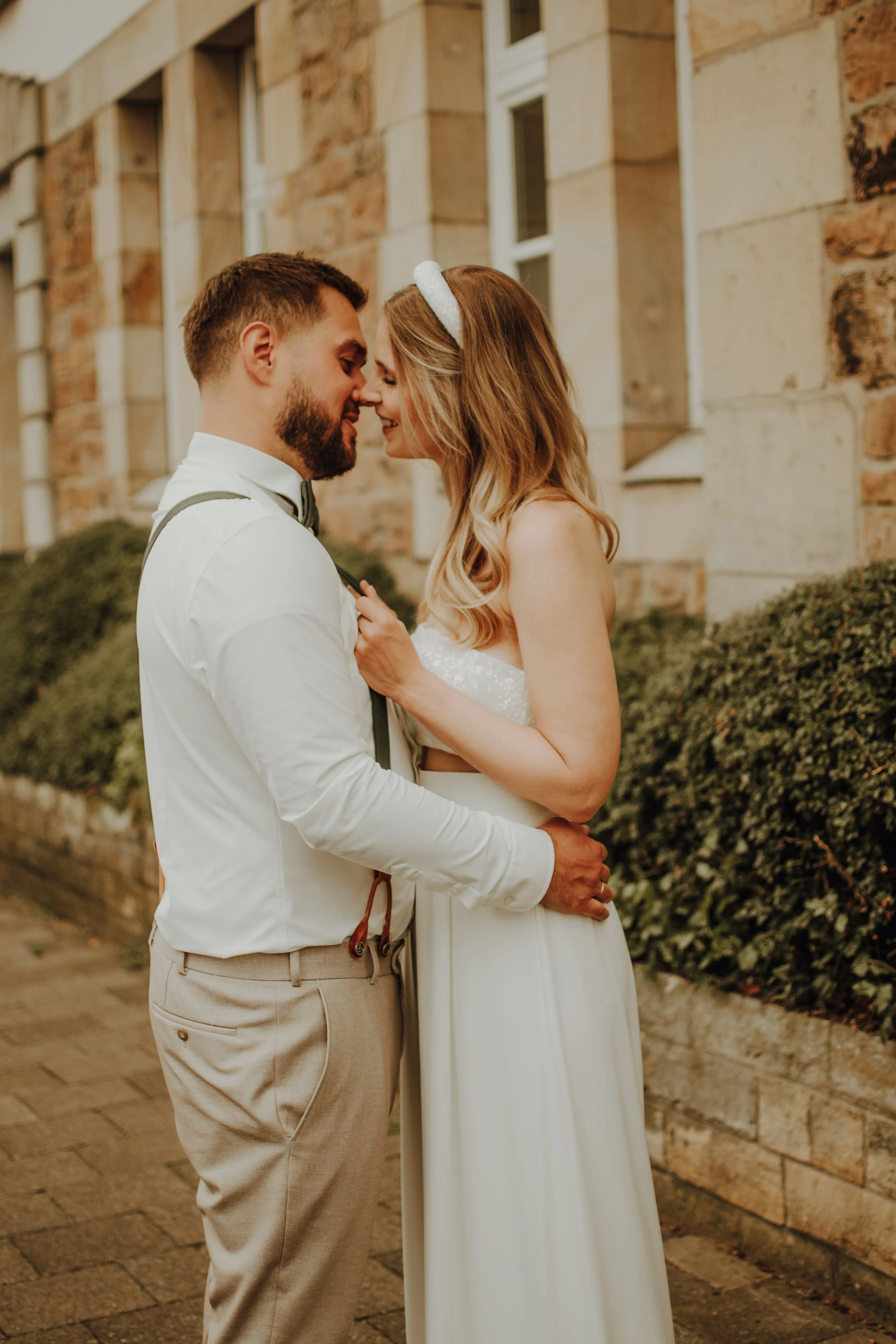 Ein verliebtes Paar bei einer Hochzeit, steht vor einer Backsteinmauer, umarmt sich und lächelt.