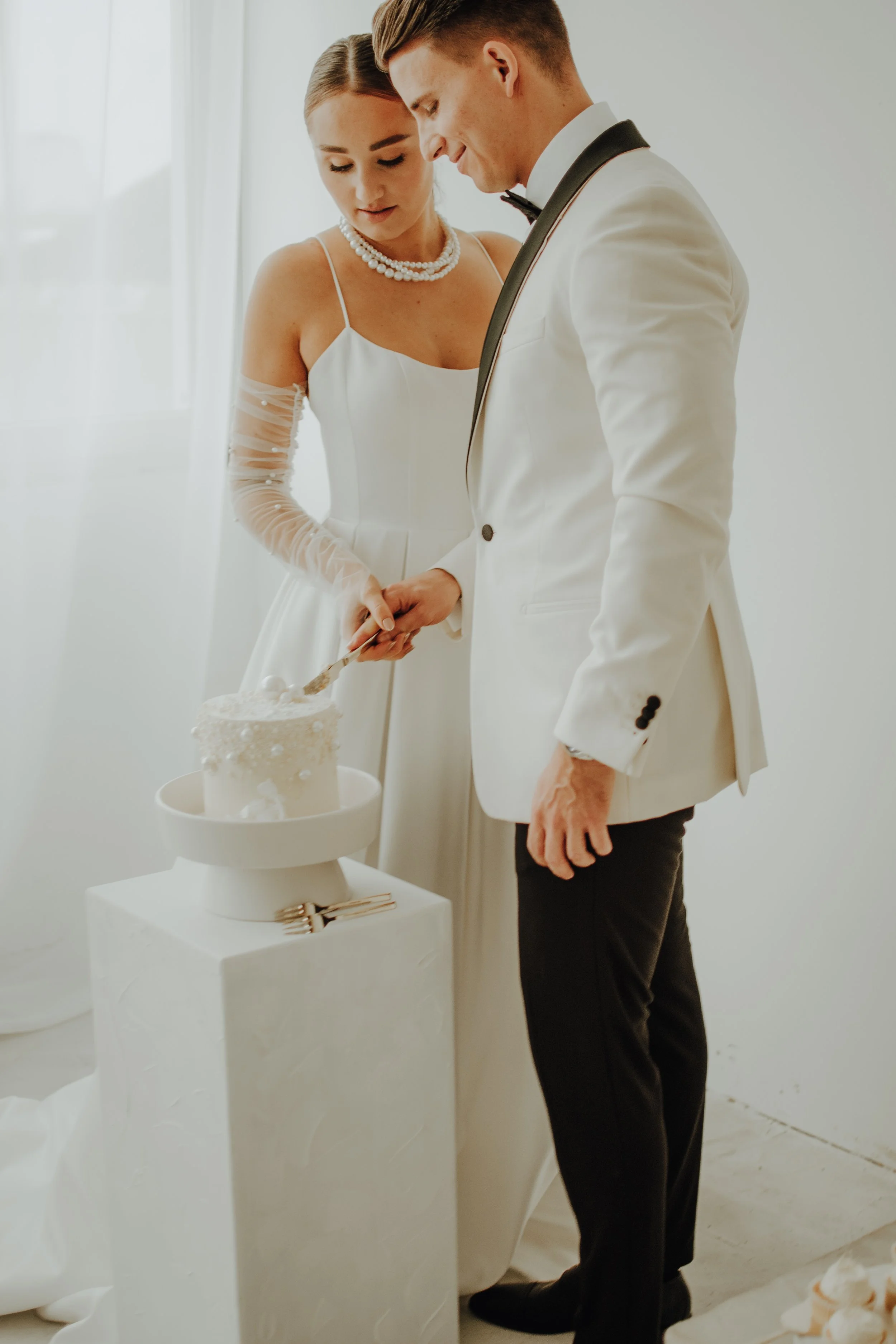 Ein Brautpaar beim Anschneiden einer Hochzeitstorte im weißen Raum.