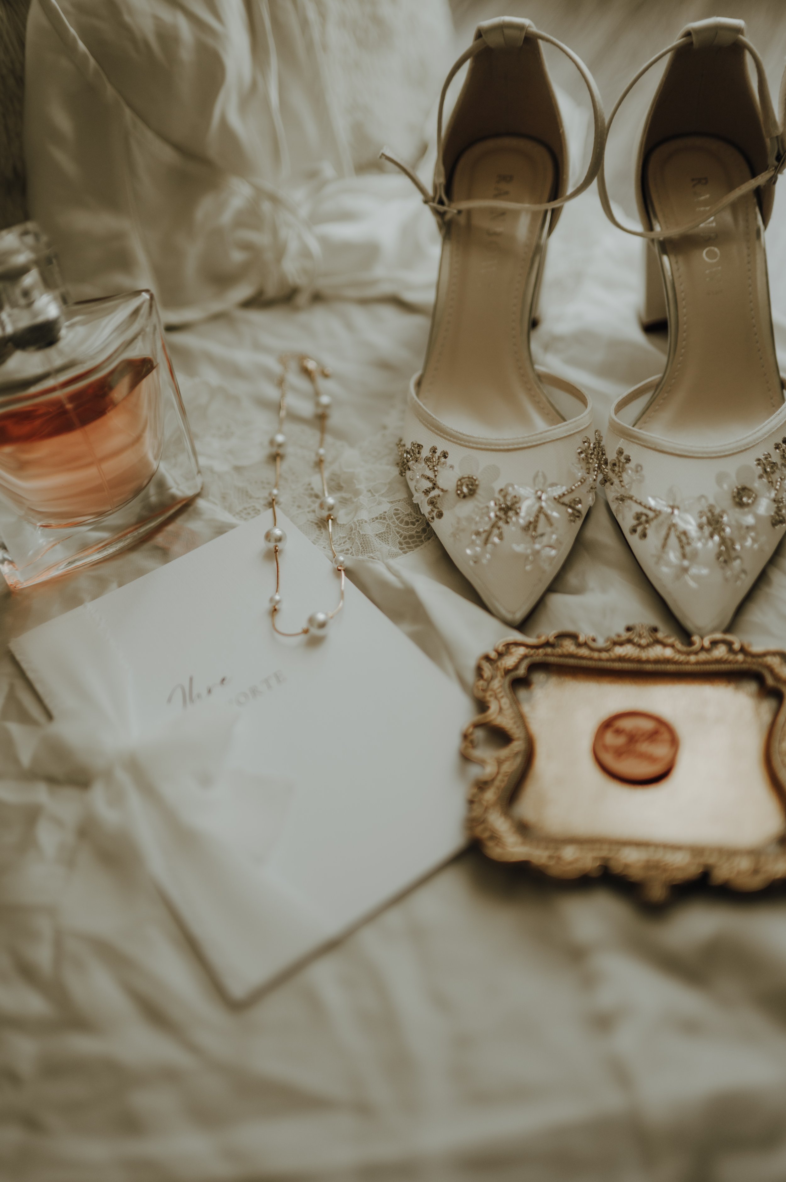 Hochzeitsschuhe, Schmuck und Parfüm auf einem cremefarbenen Stoff, vermutlich Brautaccessoires.