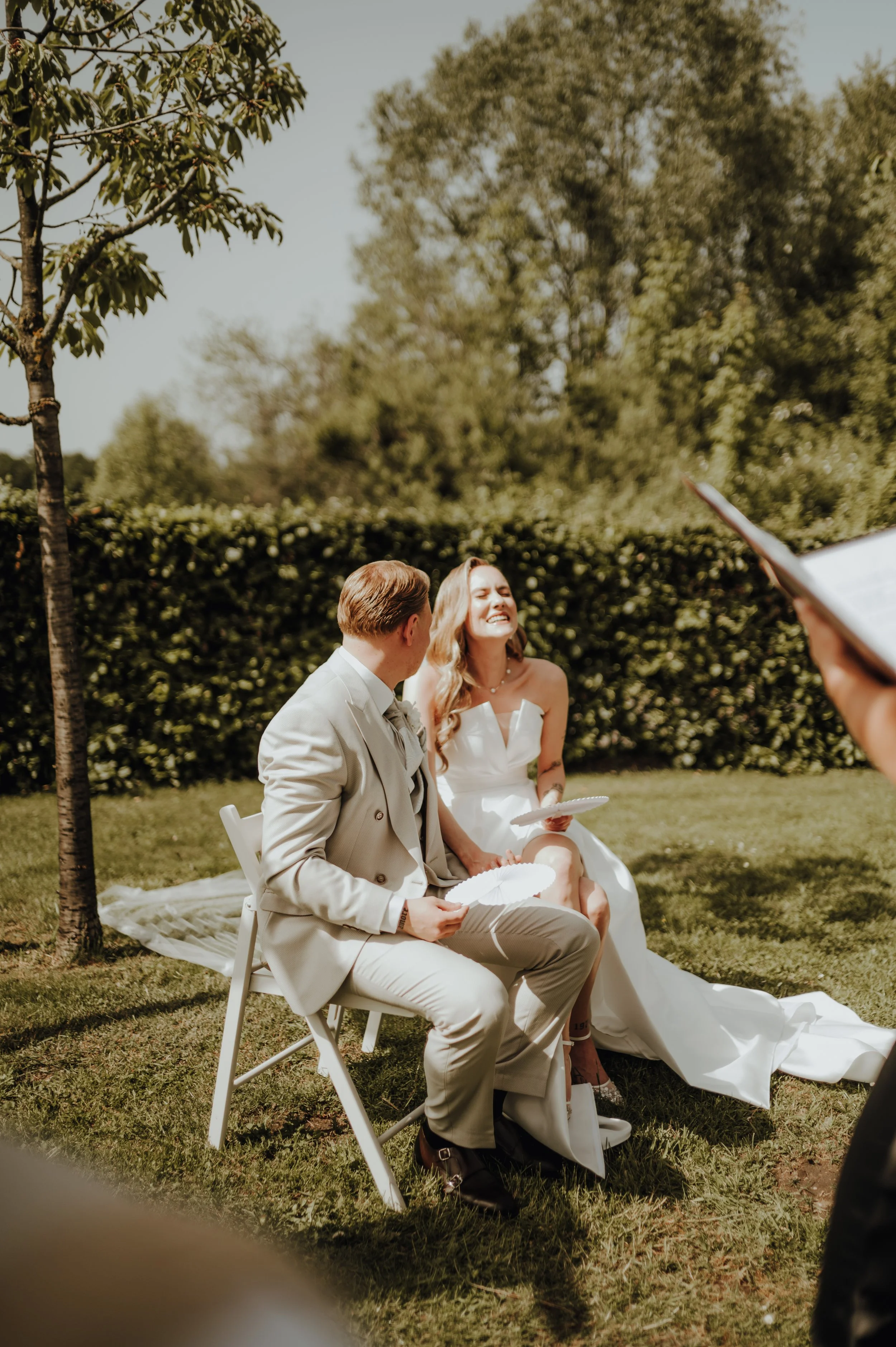 Ein Brautpaar sitzt im Garten, wobei sie lachen und sich anlächeln, während eine Person im Vordergrund eine Zeremonie mit Papierblumen durchführt.